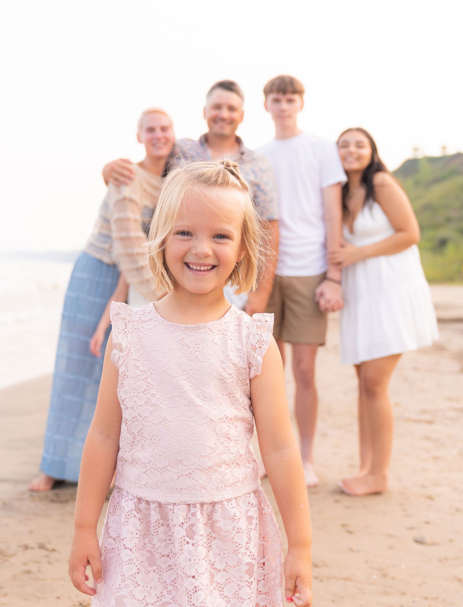 A smiling young girl in a pink dress stands in front of a group of people on a beach.