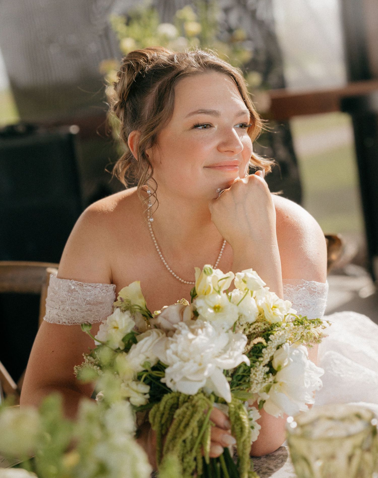 A bride in an off-shoulder lace gown holds white floral bouquet, gazing dreamily while seated outdoors.