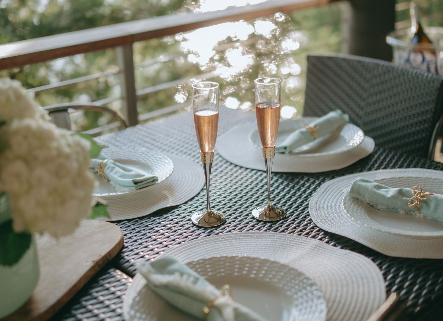 Two champagne flutes sit on a table with white plates and flowers in an outdoor setting.