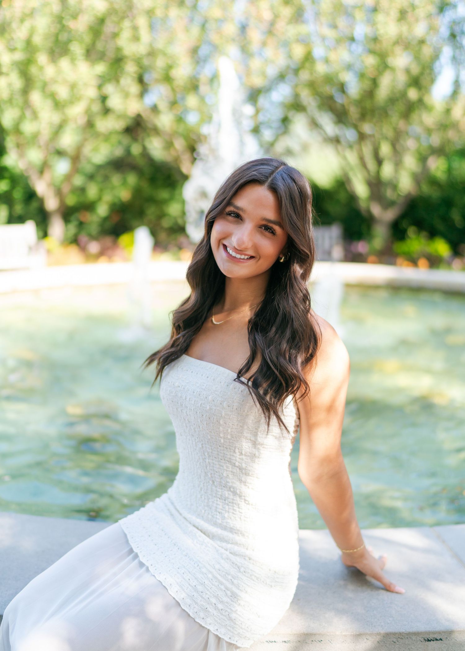 Young woman in white strapless dress smiling by a fountain in a lush garden setting.