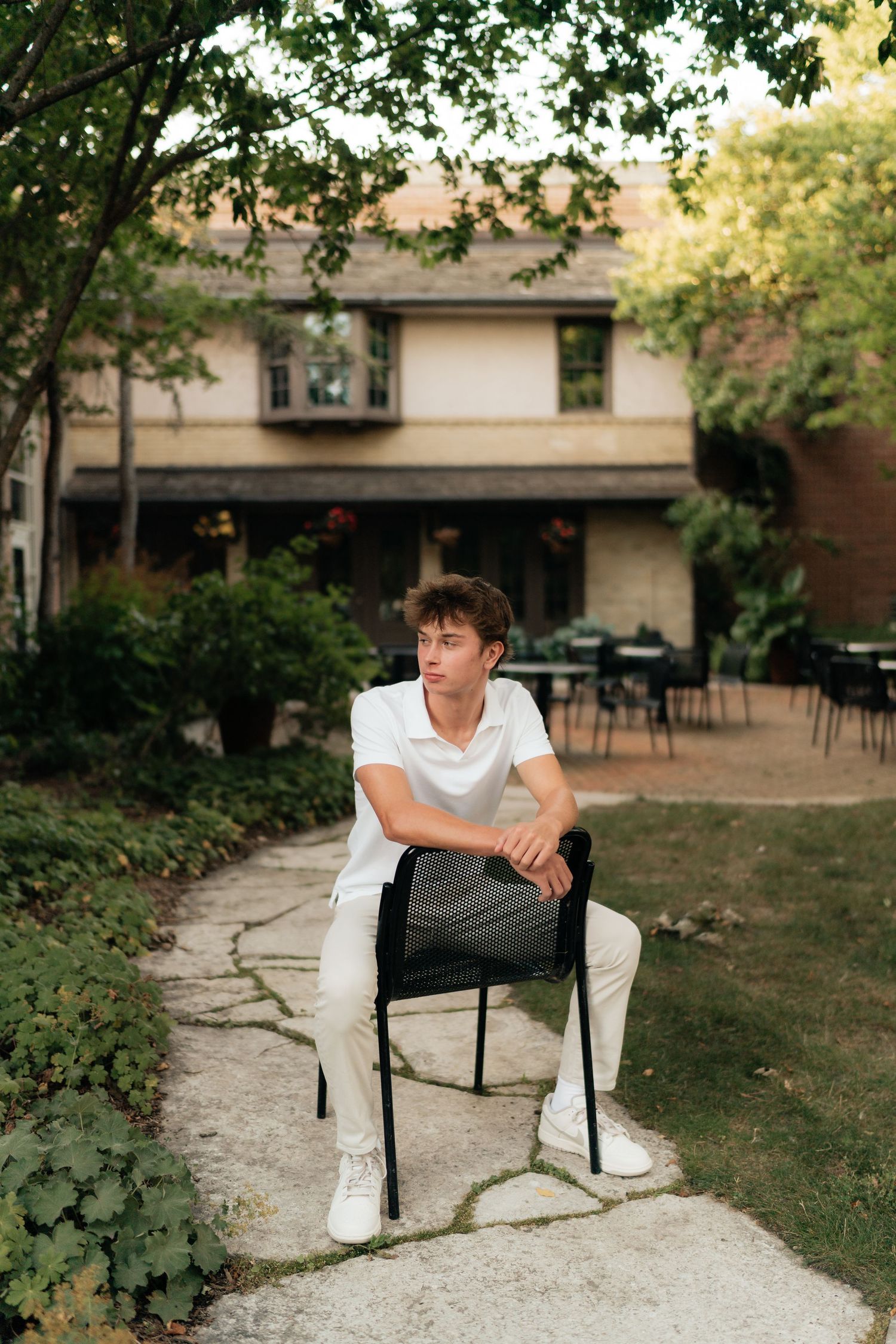 Young man in white outfit sitting on a black chair in a garden courtyard with lush greenery and building backdrop.