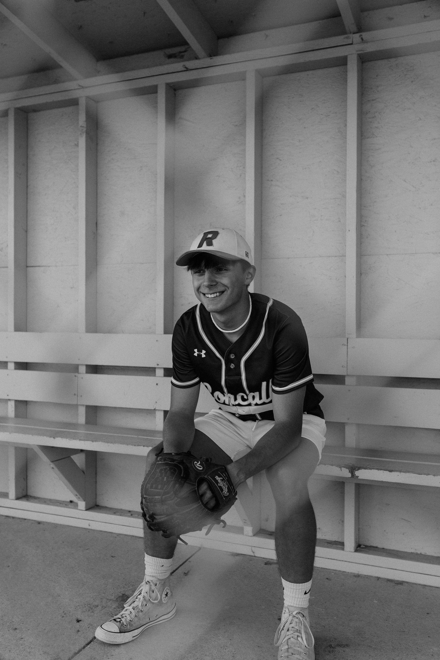 A black and white photograph of a baseball player sitting on a bench in a stadium, wearing a uniform with the letter 'R' on his cap.