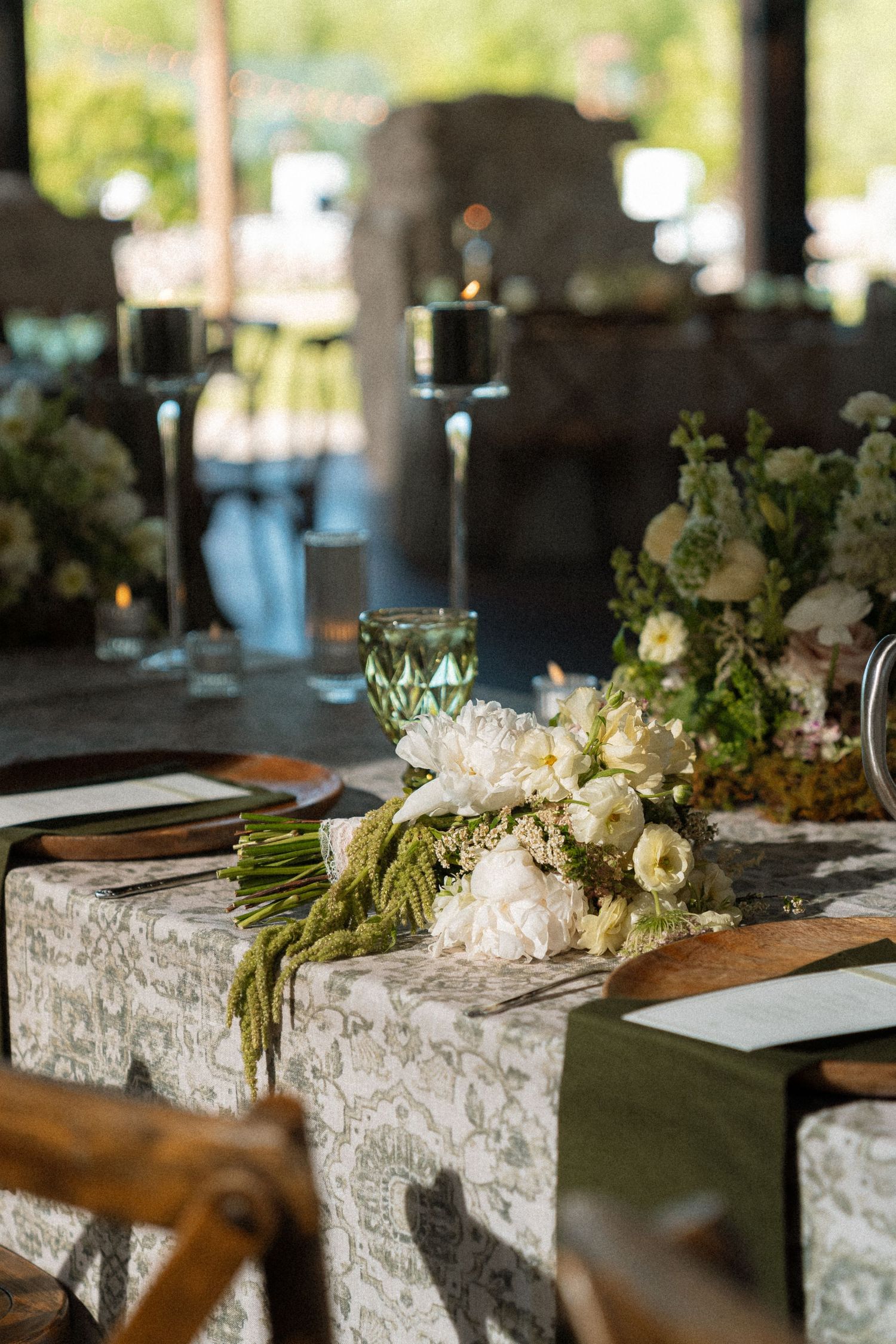 Elegant wedding table with white floral bouquet, lace runner, green glassware, and candles in an outdoor venue.