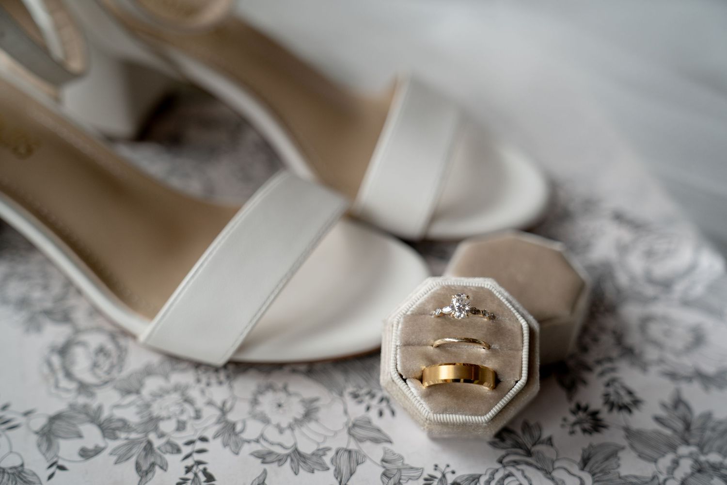 A close-up view of a pair of white high-heeled shoes with a gold ring detail on a floral lace background.