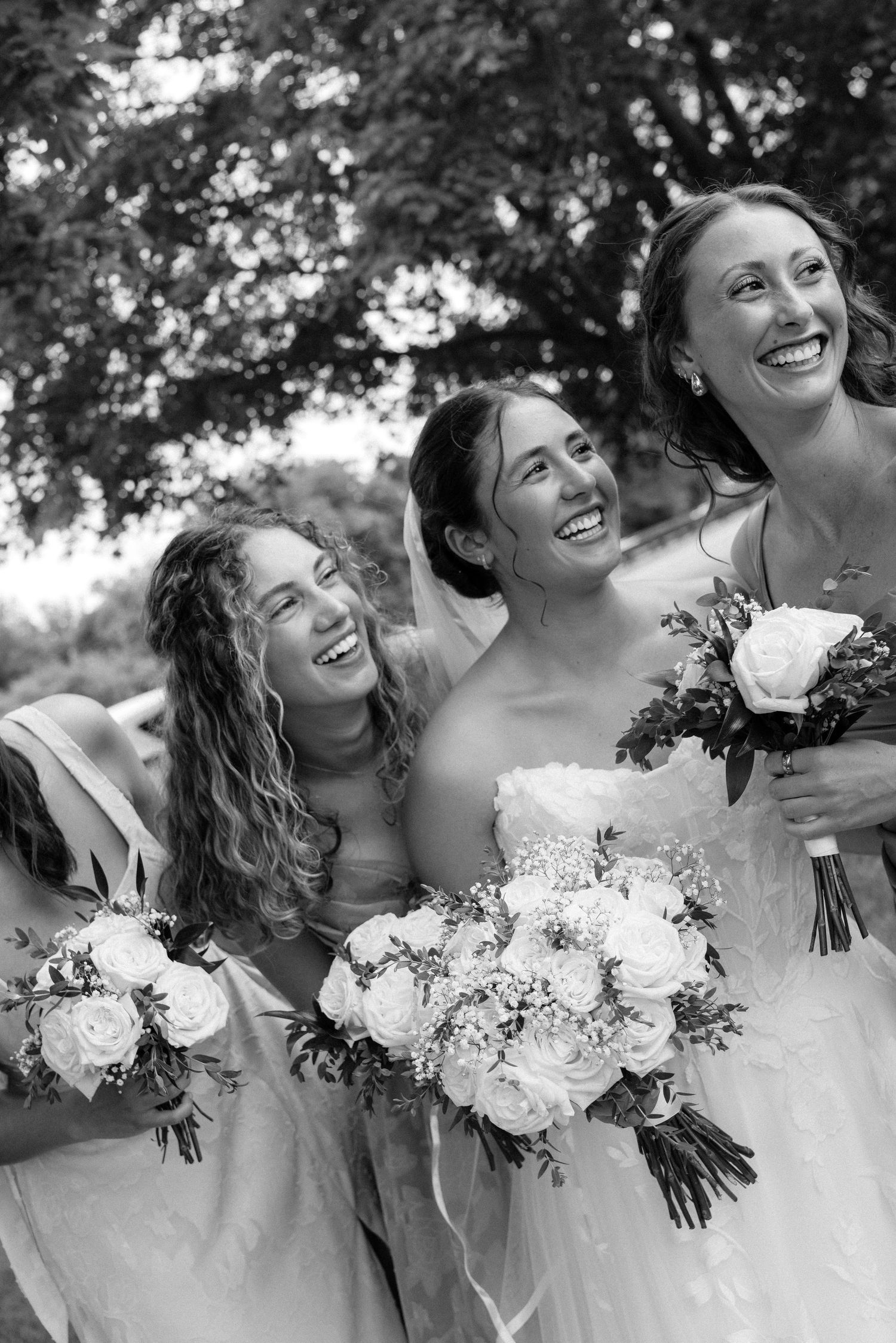 Black and white photo of a bride and two bridesmaids laughing joyfully while holding bouquets of white roses outdoors.