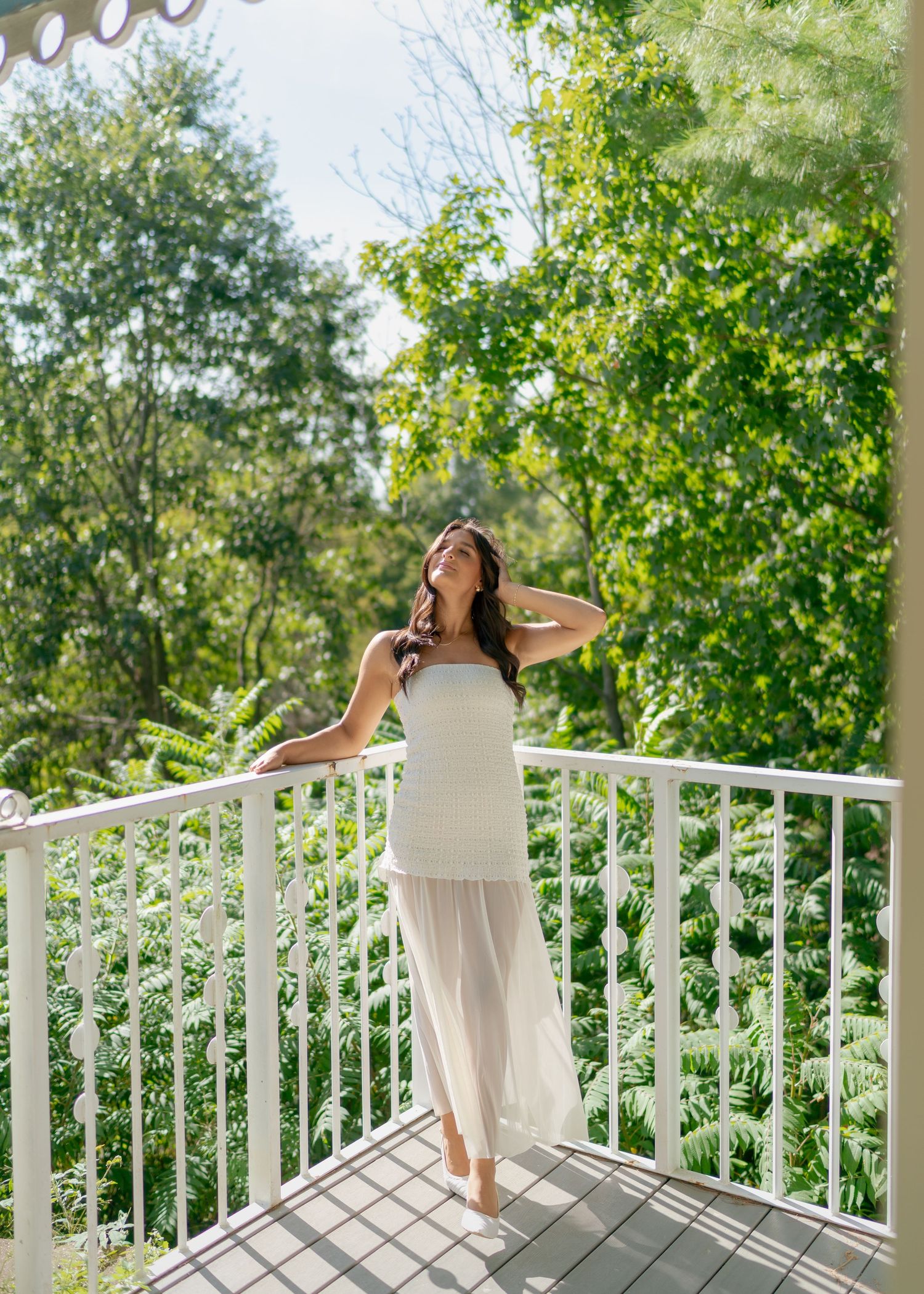 Woman in white dress standing on a white railing balcony surrounded by lush green trees on a sunny day.