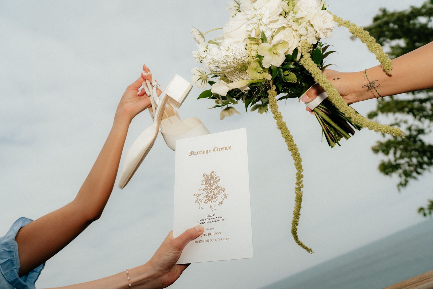 Hands holding white floral bouquet, wedding program, and bridal shoes outdoors near ocean.