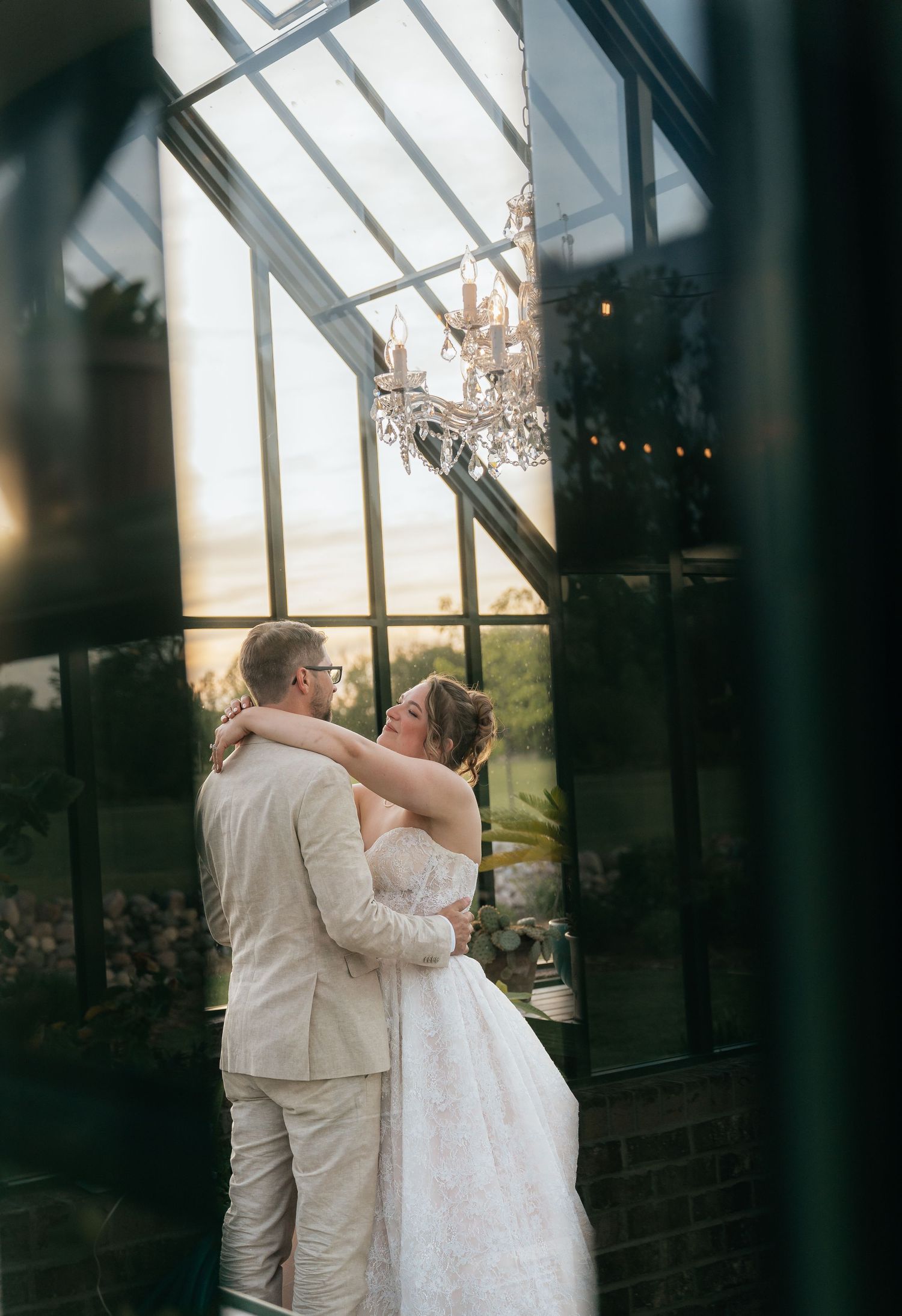 Bride and groom share romantic embrace in elegant greenhouse wedding venue with chandelier and golden sunset backdrop.