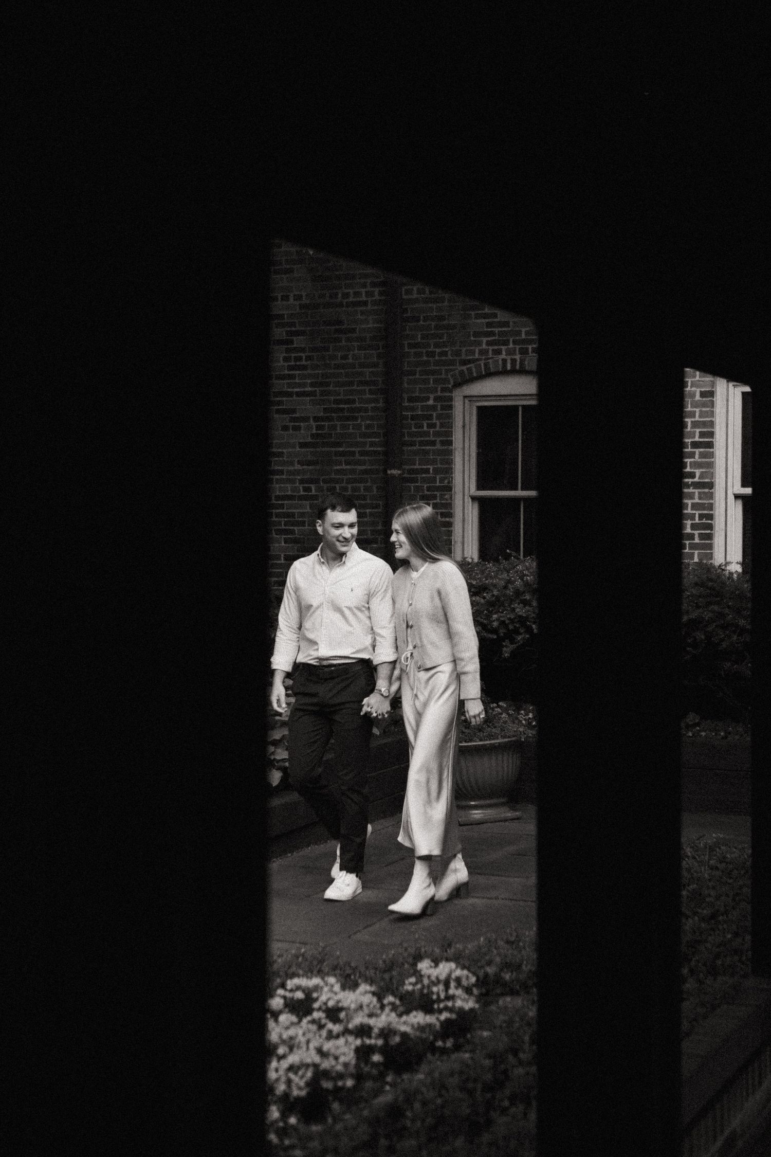 A black and white image showing two people, a man and a woman, standing together in front of a brick building.