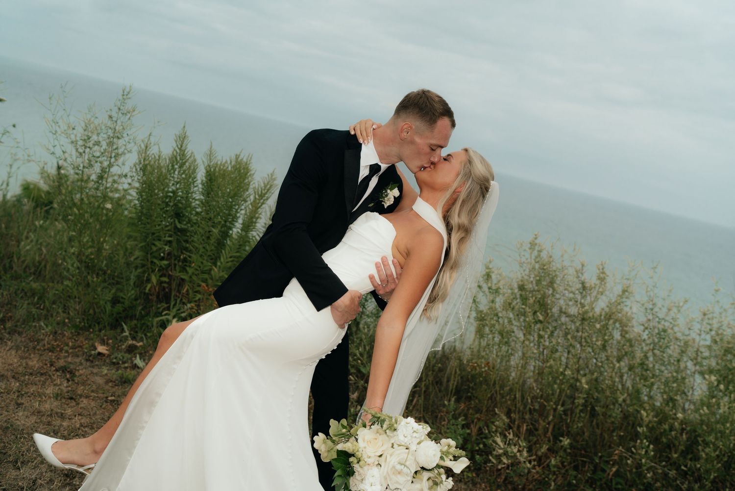 Bride and groom share a romantic dip kiss outdoors with ocean view backdrop on their wedding day.
