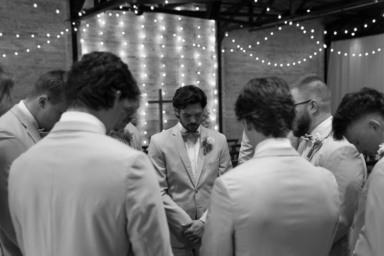 A group of men in white suits gathered together, with string lights and a backdrop visible in the background.