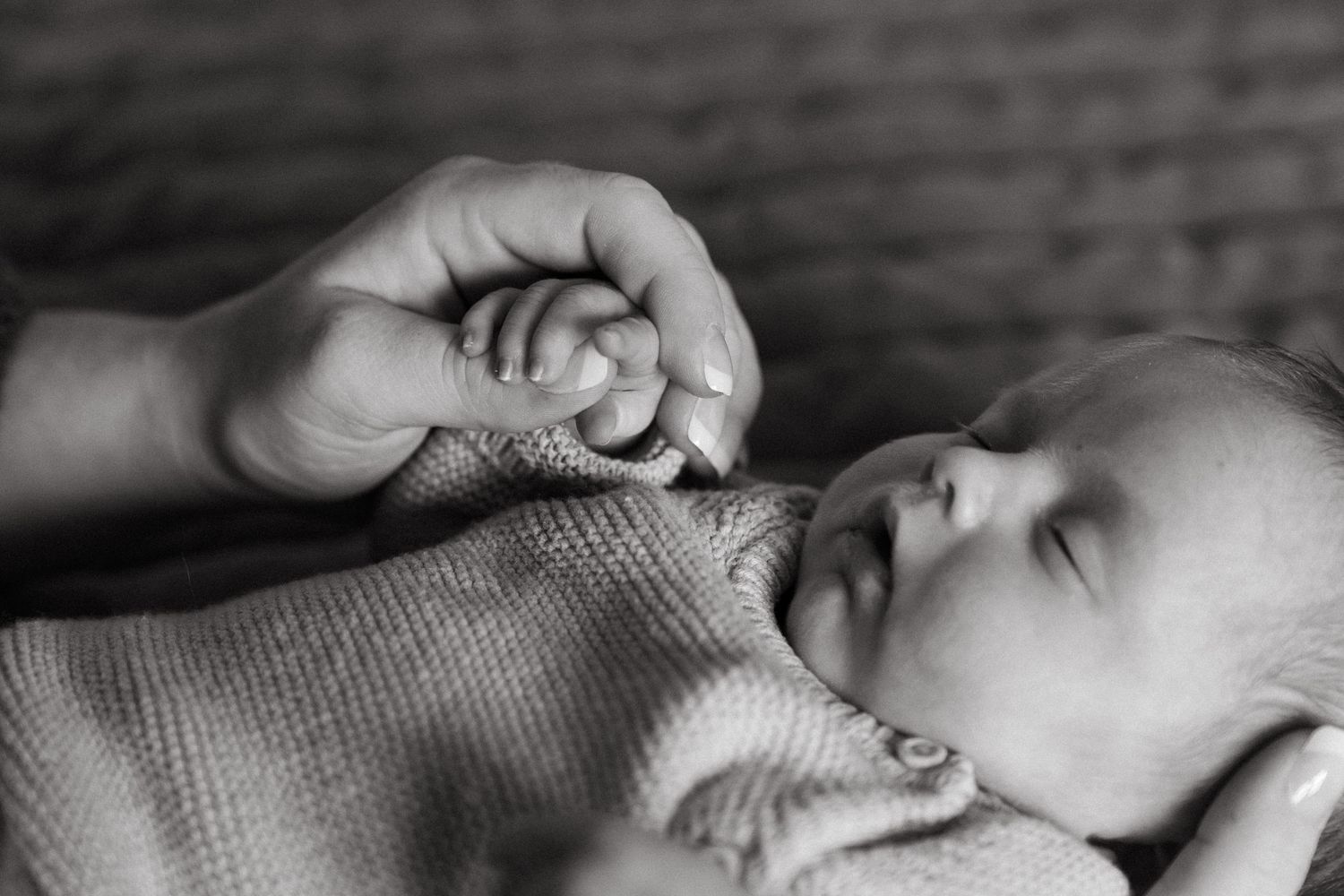 A black and white close-up image of a sleeping newborn baby's face with a hand gently touching the baby's cheek.