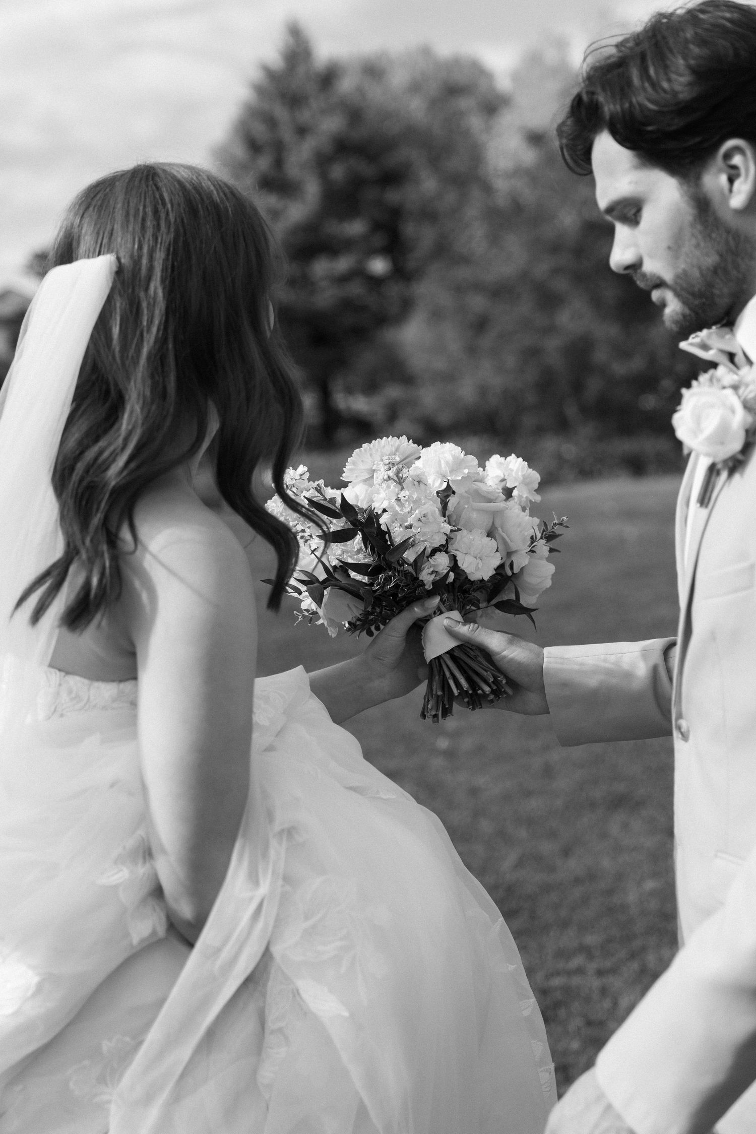 A black and white image of a bride and groom embracing and holding a bouquet of flowers in a garden setting.