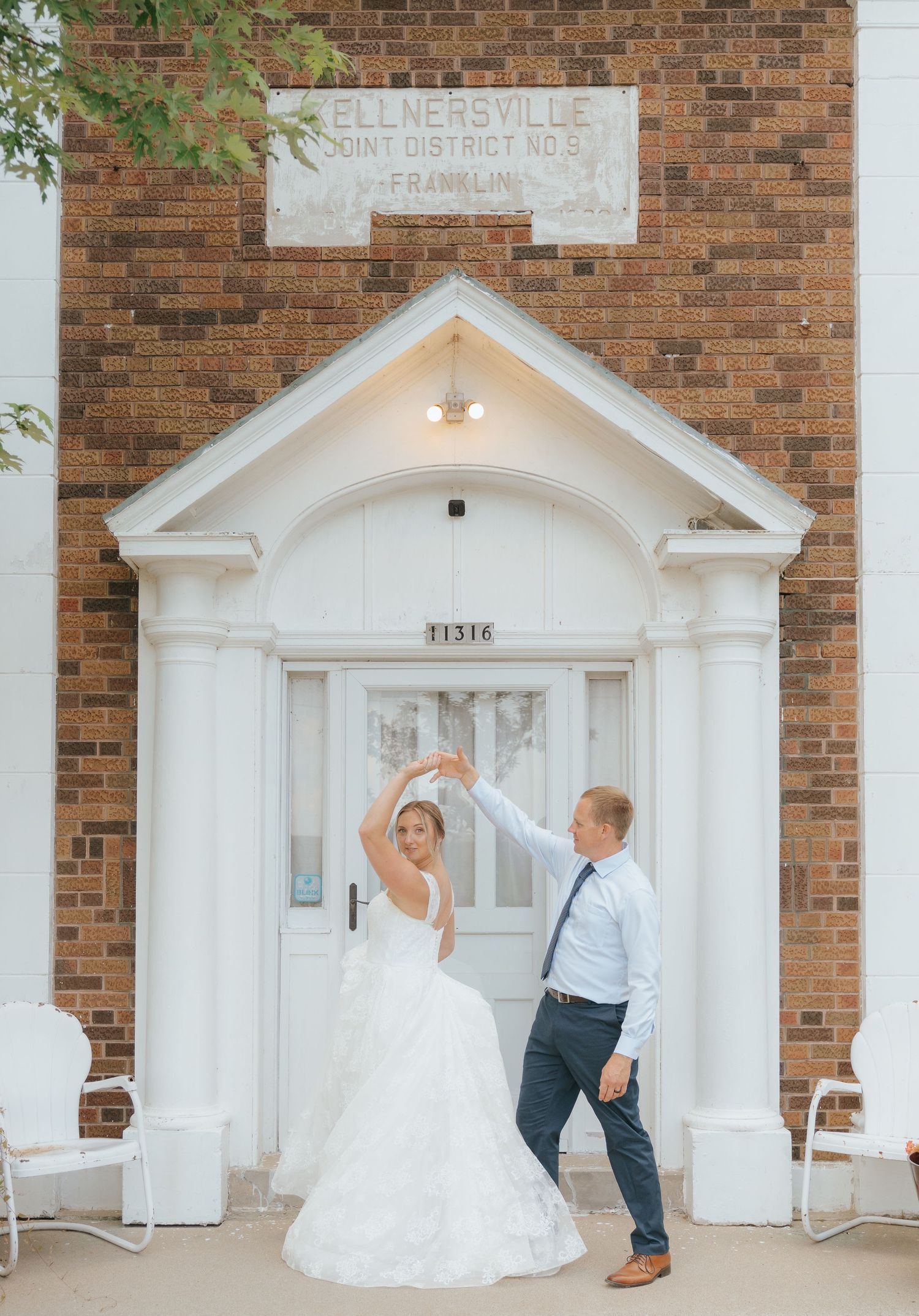 A bride and groom standing in a white archway entrance of a brick building, with a sign that says 'Kellersville' above them.
