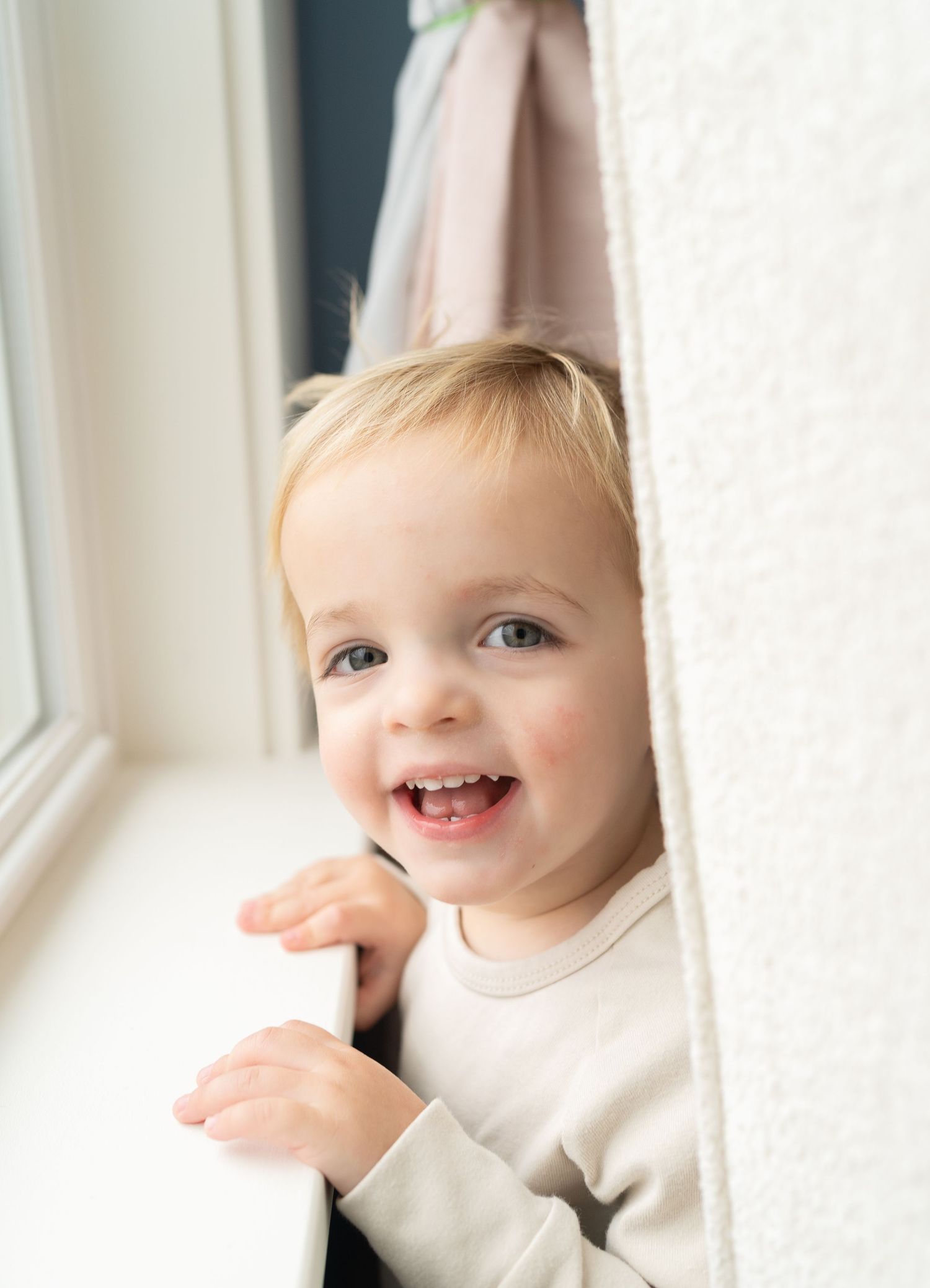 A smiling baby boy with blond hair looking out a window.