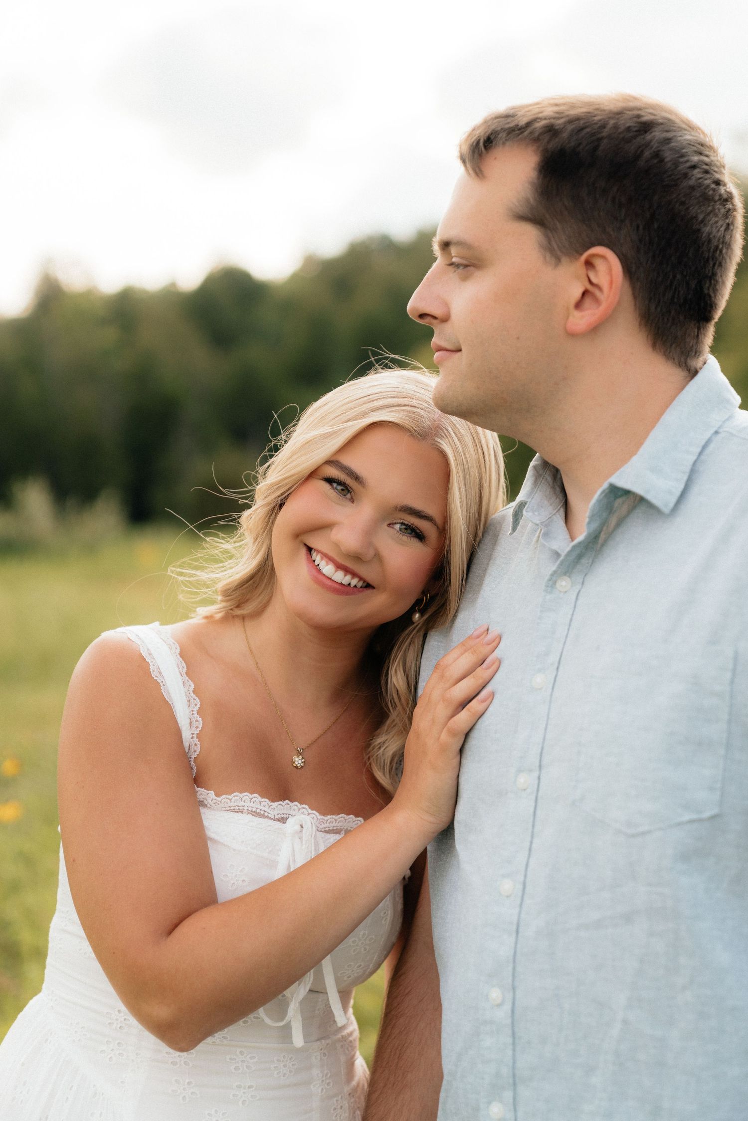 A smiling blonde woman in a white dress leans against a man in a blue shirt in a sunny green field.