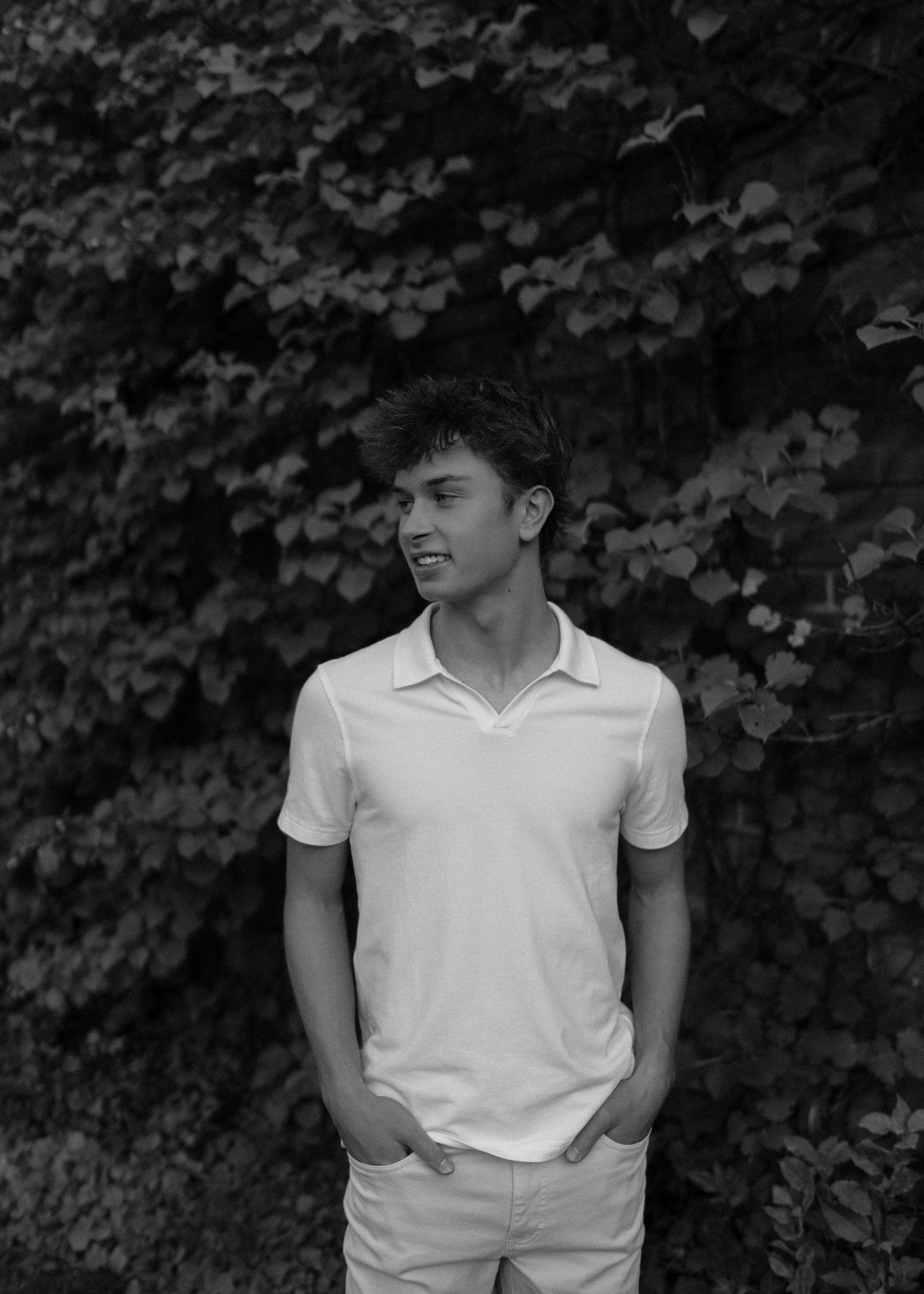 Young man in white polo shirt standing outdoors against lush ivy wall, black and white portrait photo.