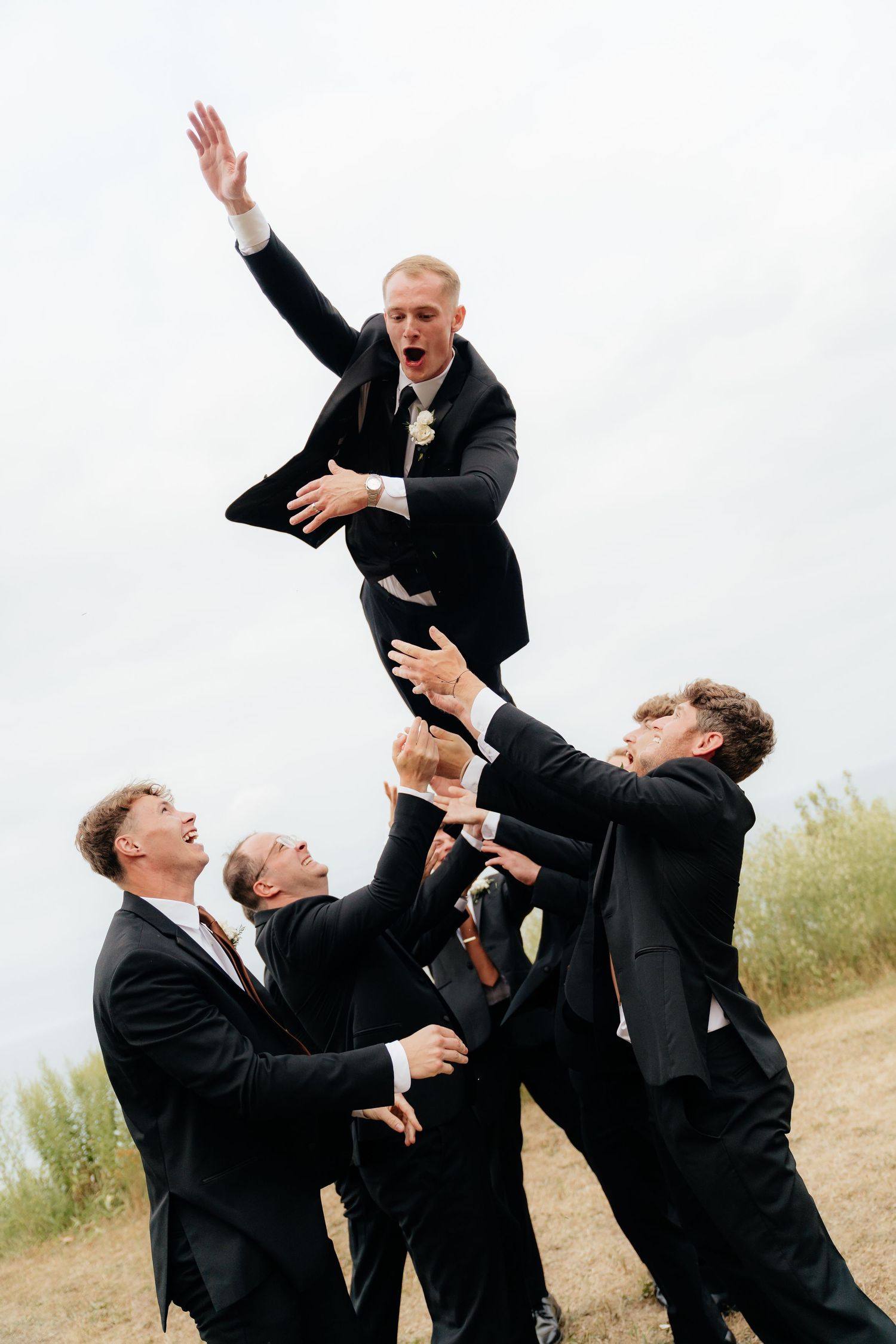 Groomsmen in black suits toss the groom into the air in a joyful wedding celebration outdoors.