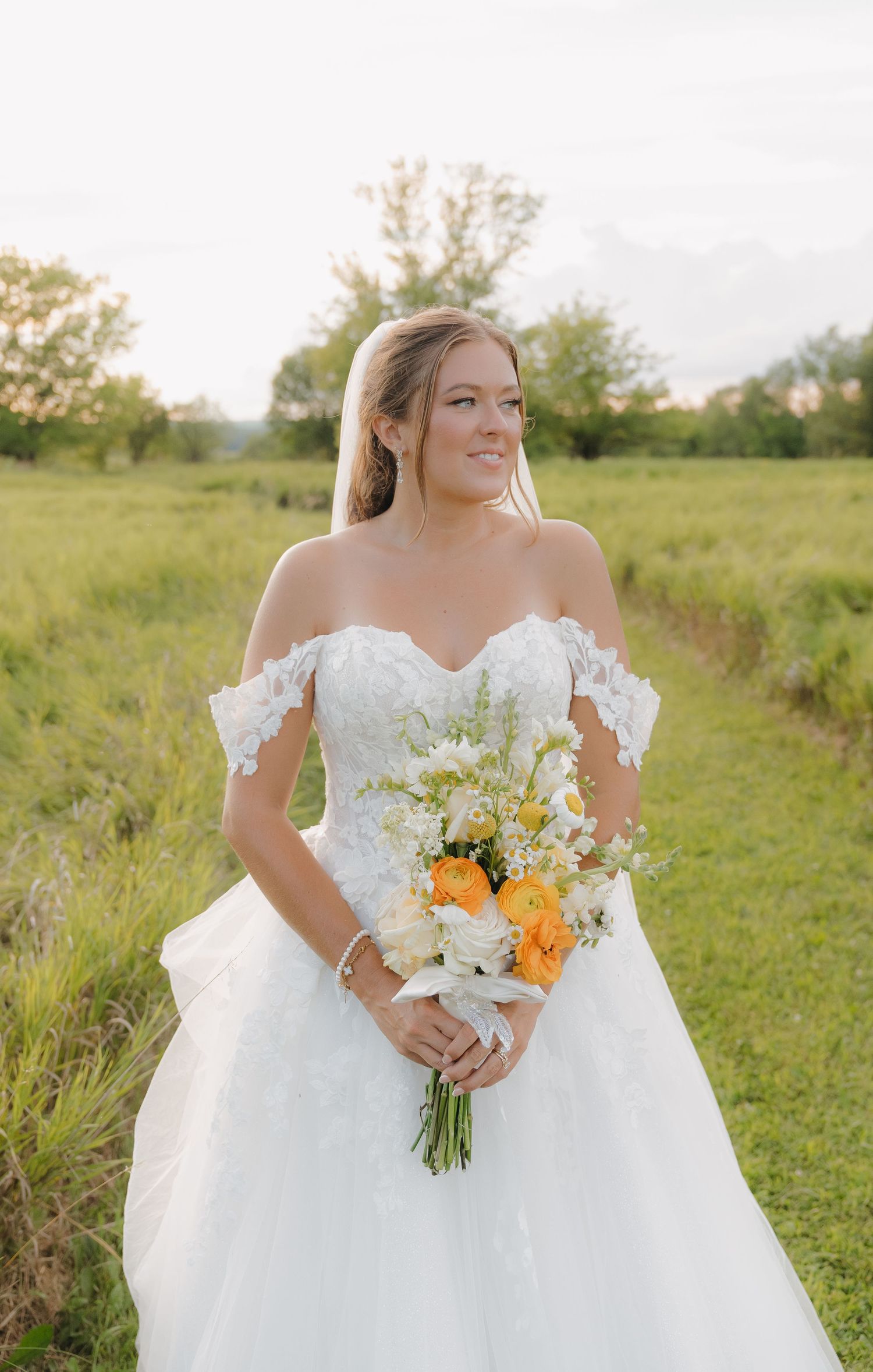 A bride in a white wedding dress stands in a grassy field, holding a bouquet of flowers and smiling at the camera.