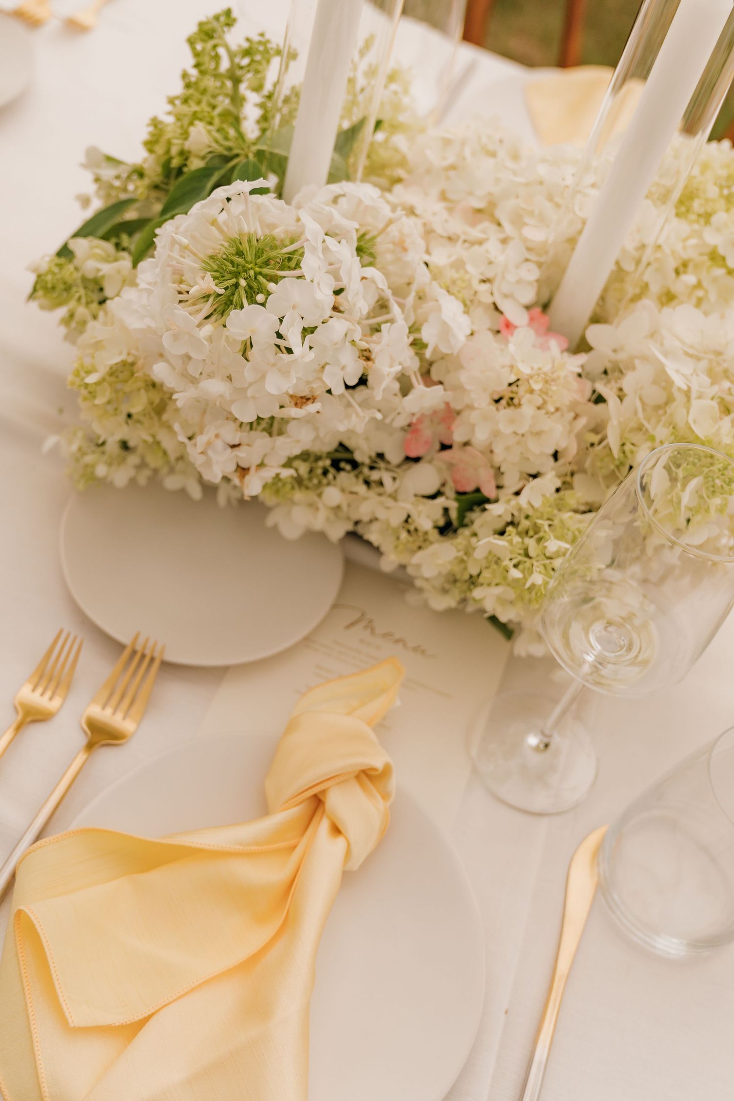 A table setting with a vase of white and green flowers, yellow napkins, and silverware on a white tablecloth.