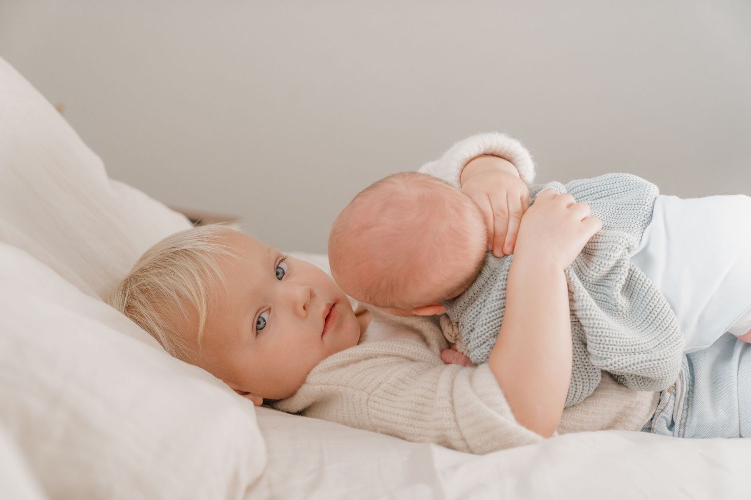 A baby lying on a white blanket, holding a peach-colored toy and wearing a gray outfit.