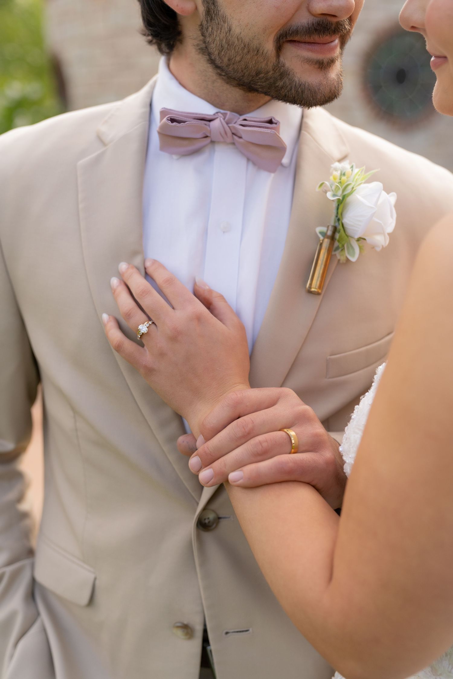 A man in a beige suit and bowtie holds a woman's hand, with a boutonnière on his lapel.