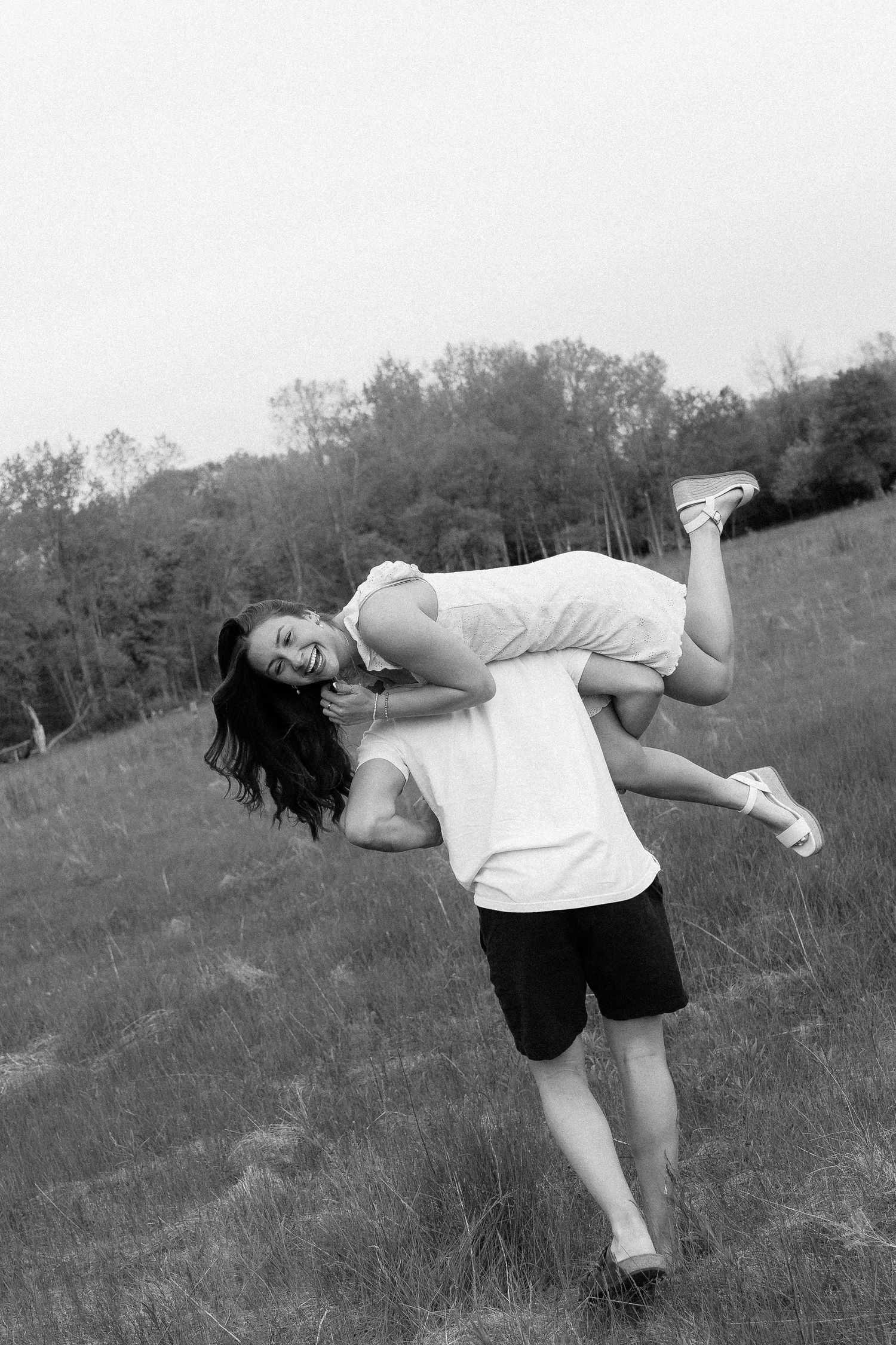 Black and white photo of a man lifting a laughing woman outdoors in a grassy field with trees in the background.