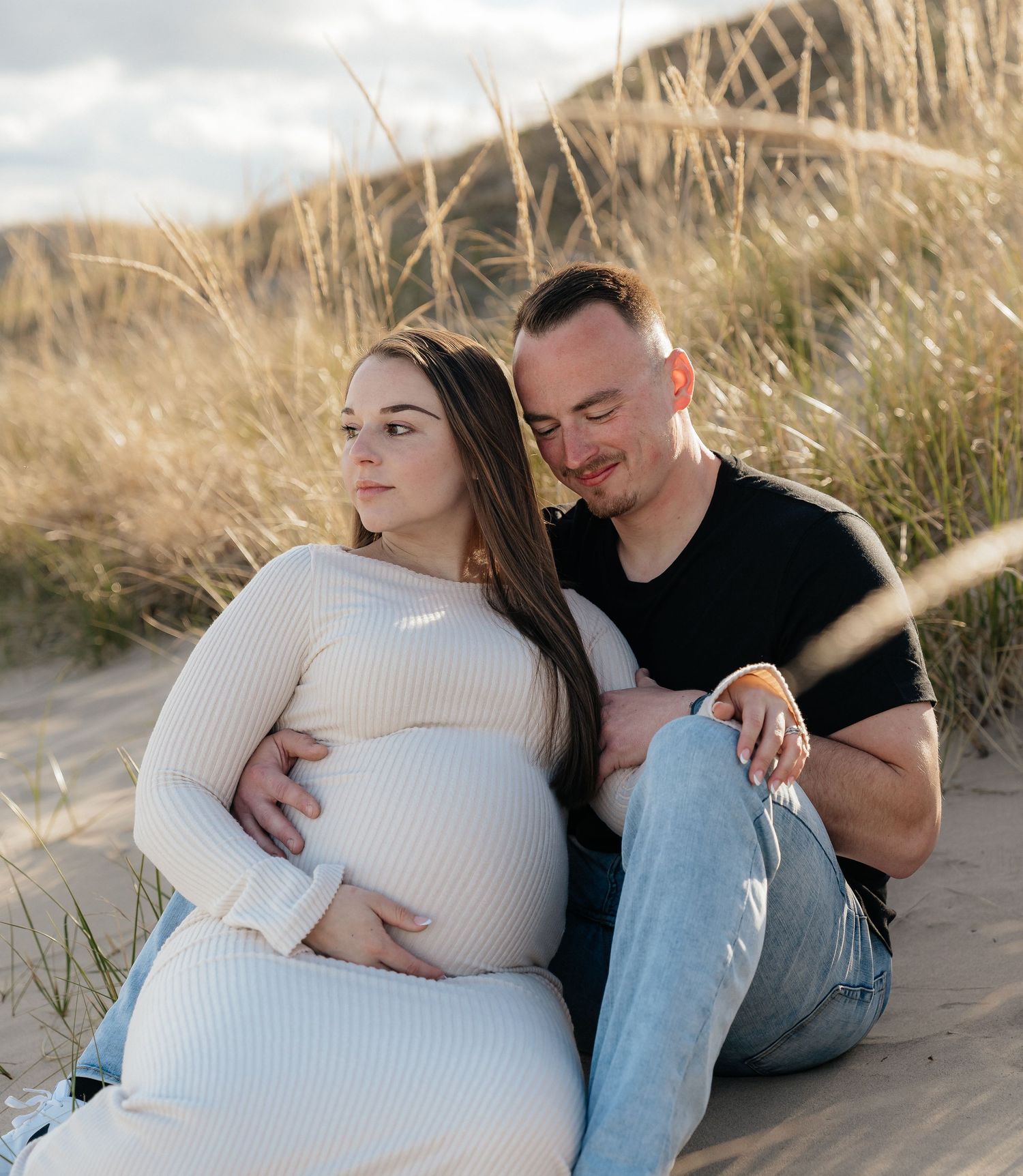 Couple sitting on sand dunes, man embracing pregnant woman in white dress during maternity photo shoot.