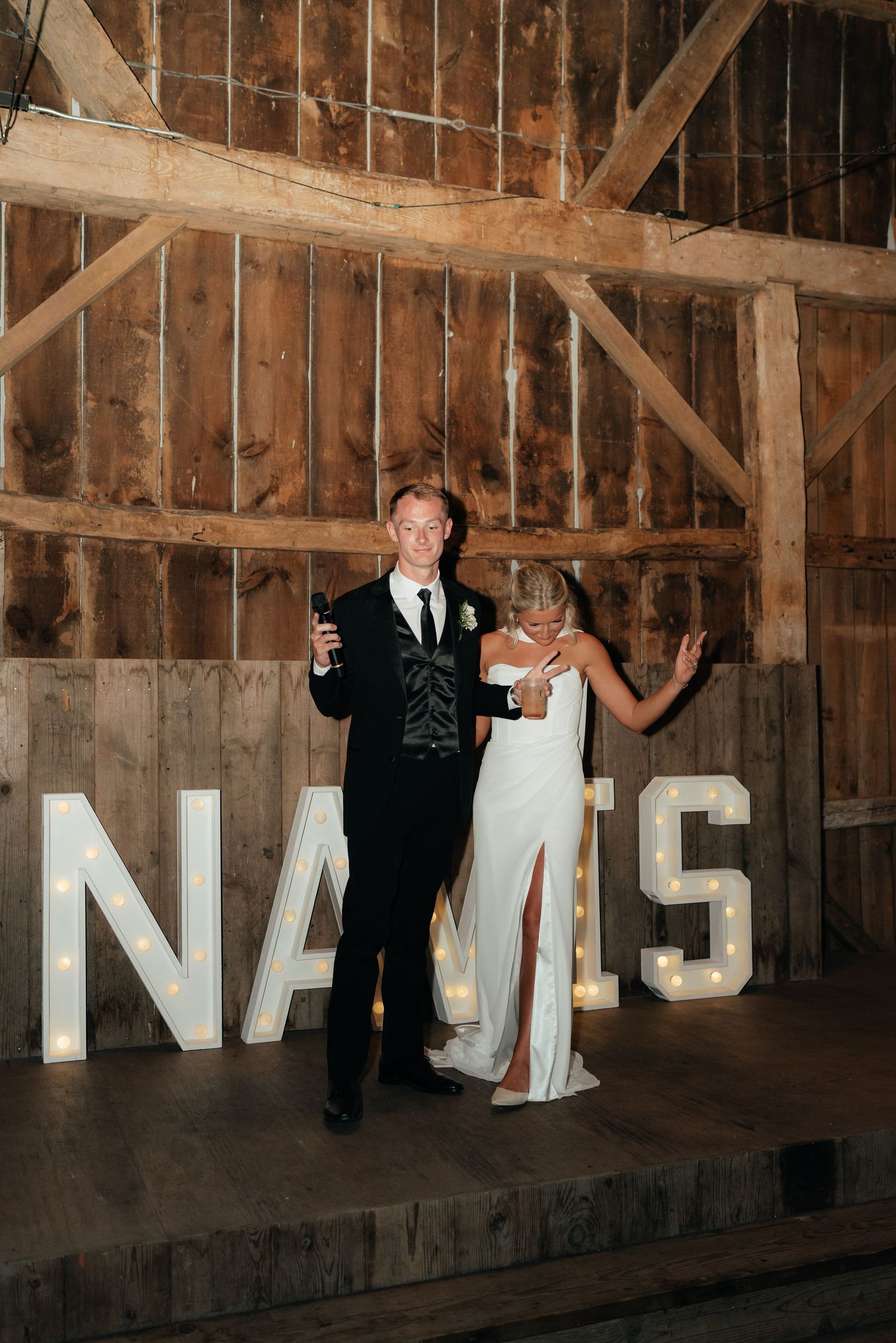 Bride and groom posing in front of marquee letter lights inside a rustic barn wedding venue.