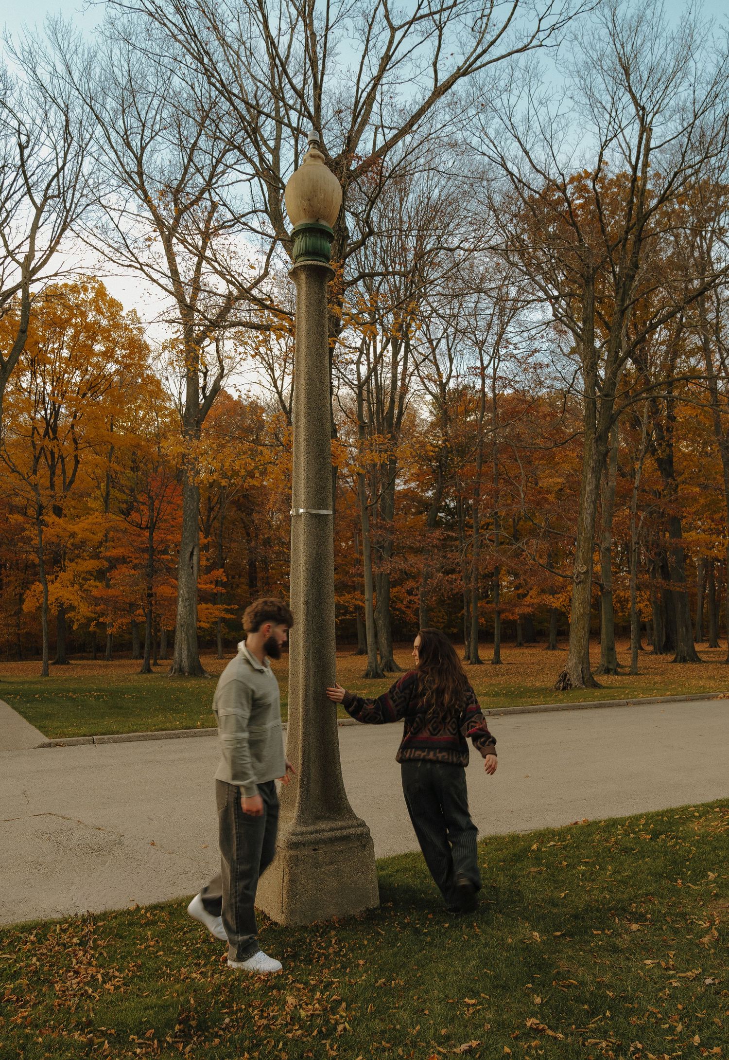 A pair of people standing near a lamp post in a park with colorful autumn trees in the background.