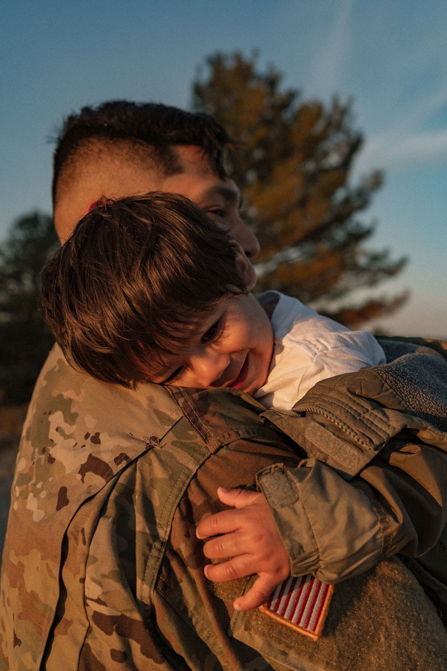 A soldier in US Army camouflage uniform holds a smiling child during a heartfelt reunion at sunset.