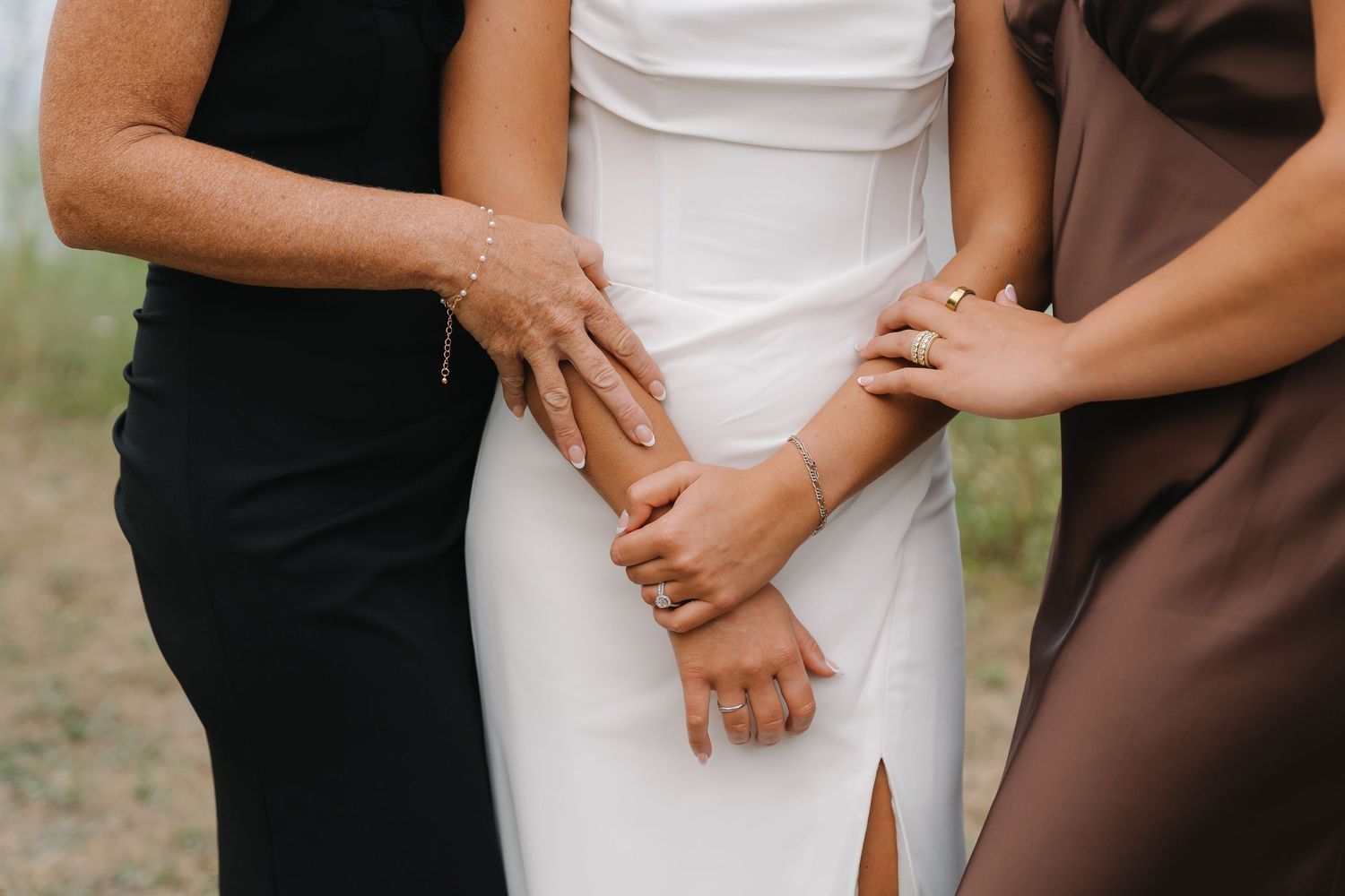 Bride in white wedding dress flanked by two women in black and brown dresses, hands clasped together.