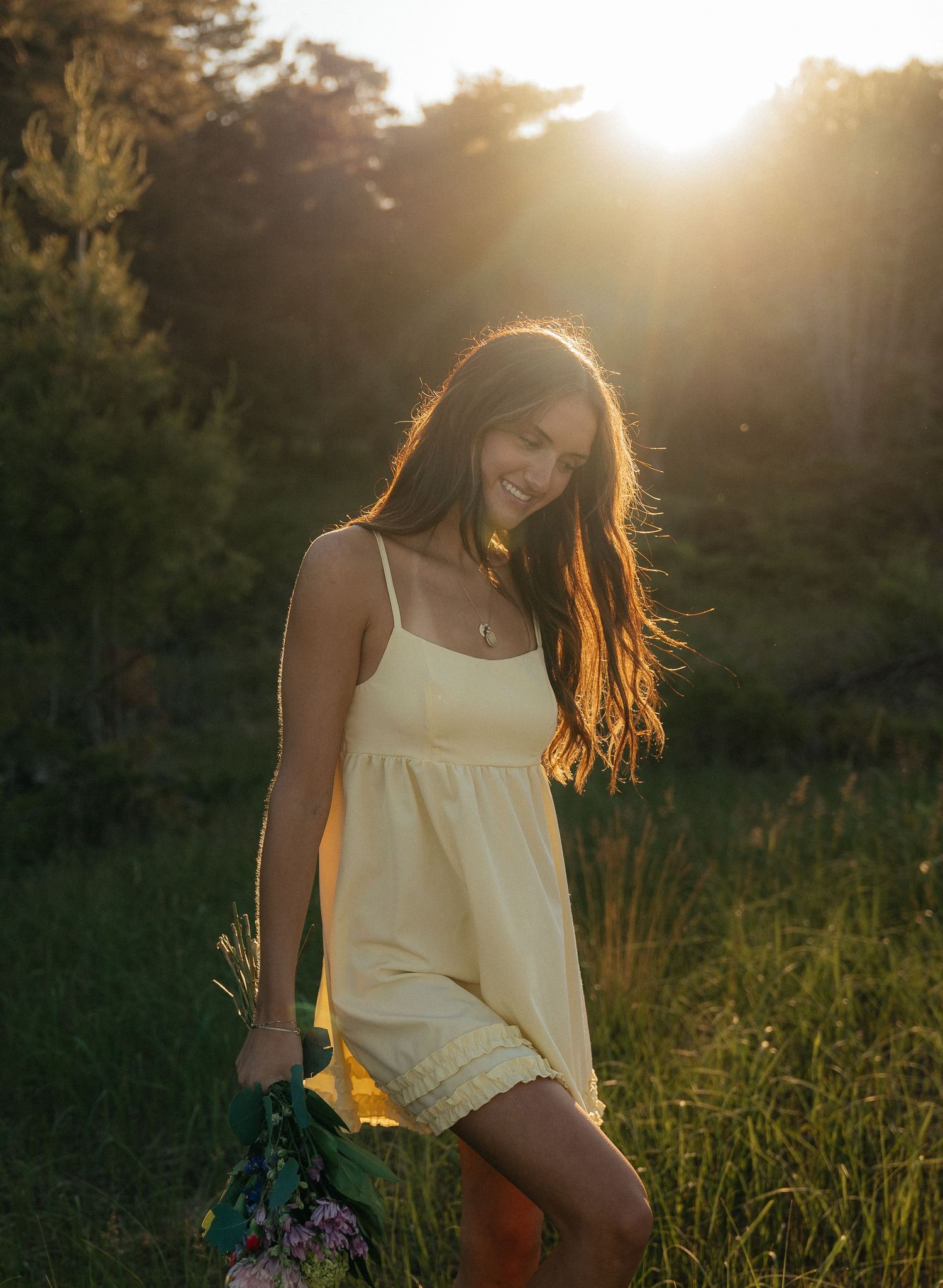 Young woman in yellow sundress smiling in golden sunset light surrounded by lush green trees and grass.