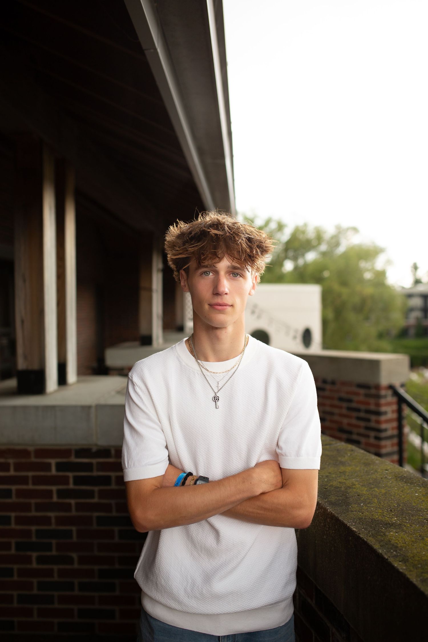 A young man with curly hair stands in front of a brick building, with his arms crossed, looking directly at the camera.