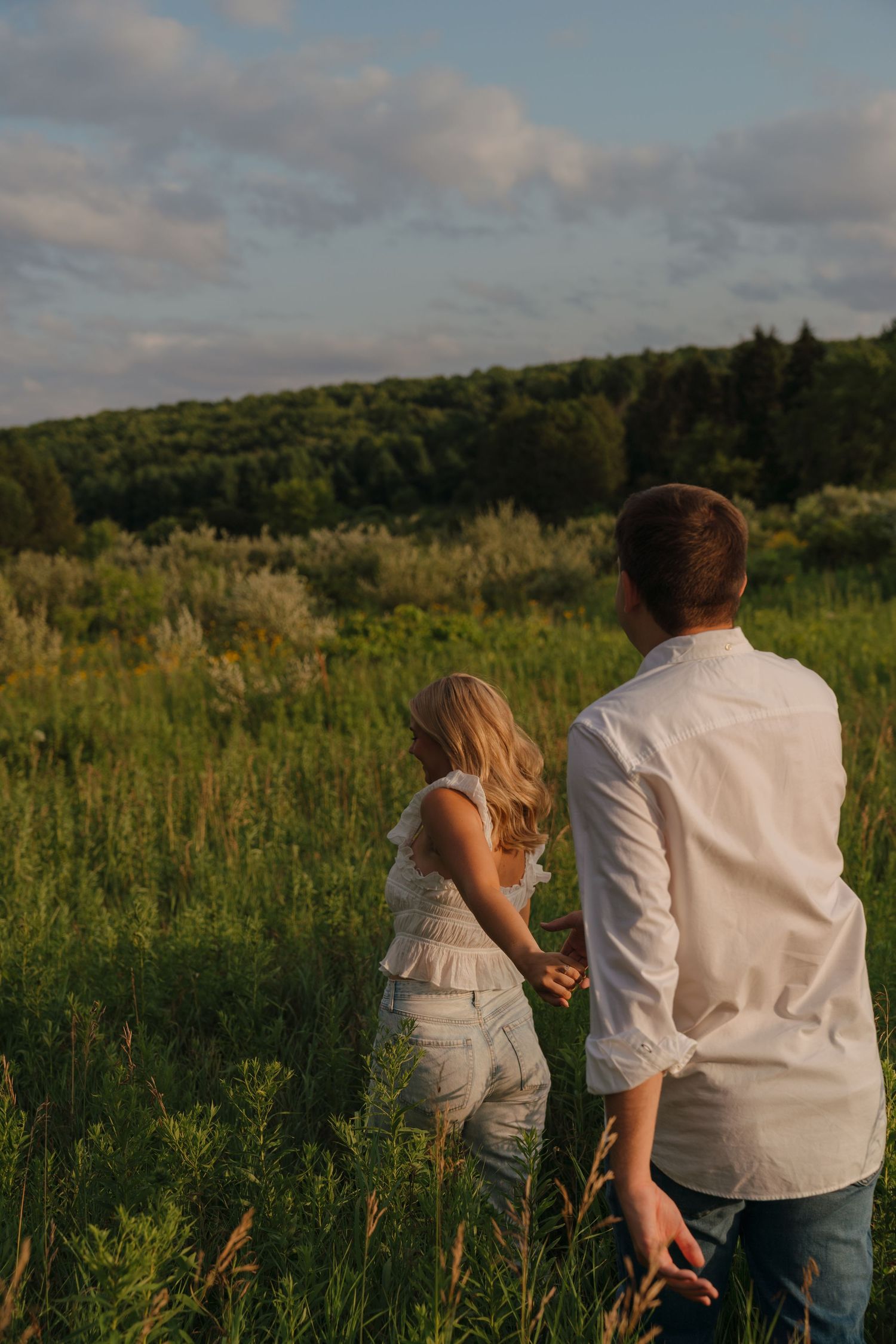 Couple holding hands walking through a lush green meadow at golden hour with forest in the background.