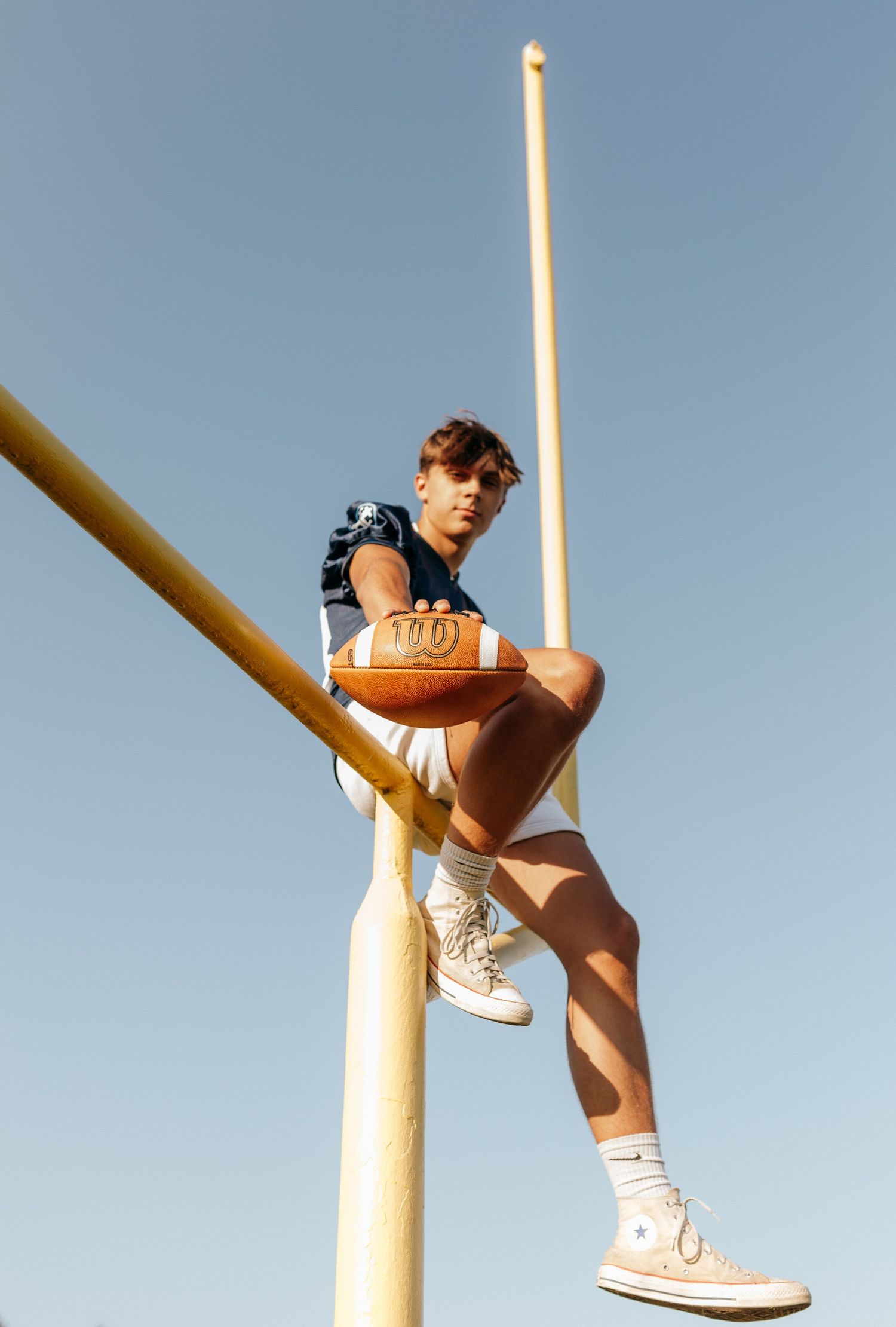 A person sitting on a pole against a clear blue sky, wearing casual clothing and holding a basketball.