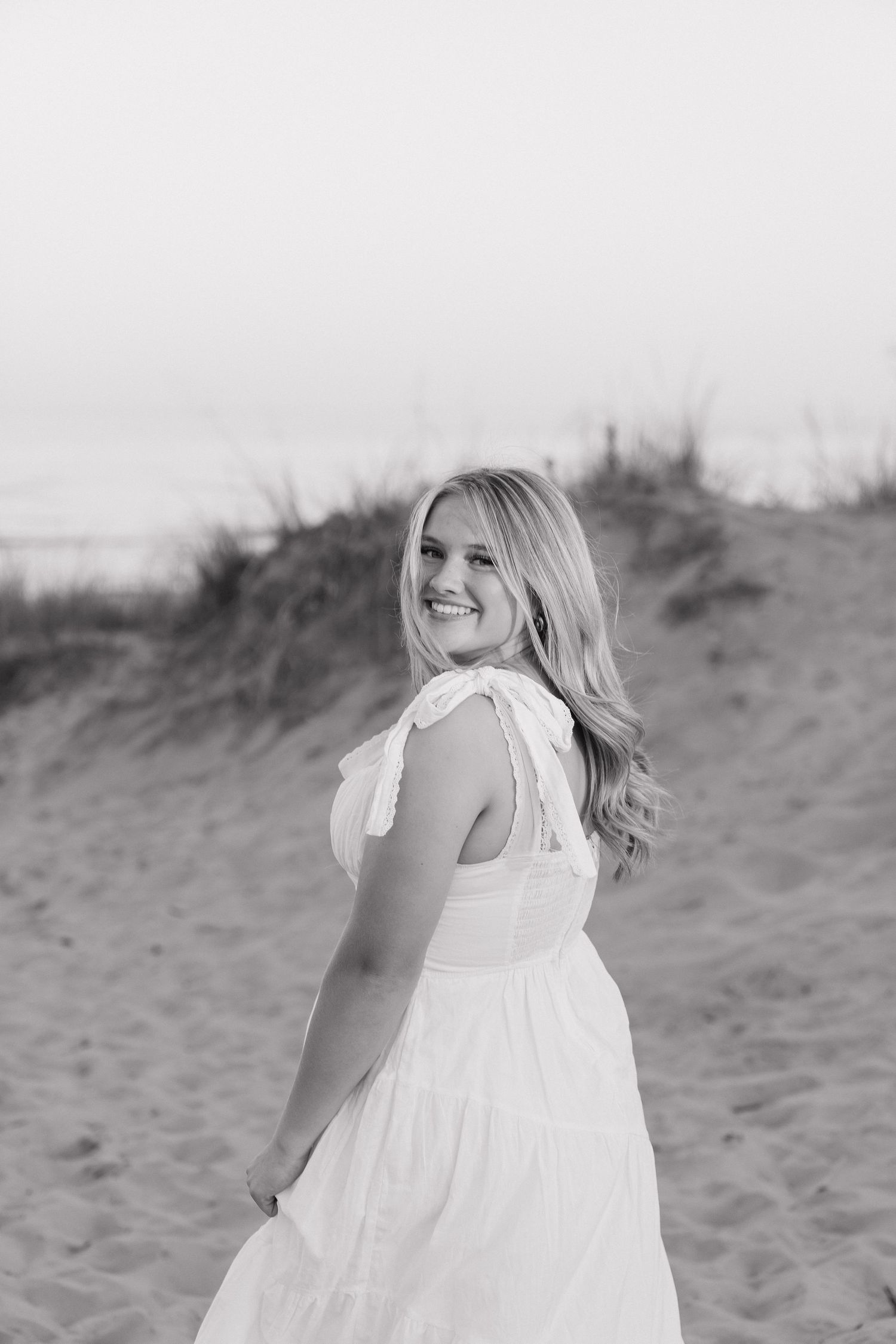 A young woman with long blonde hair wearing a white dress stands in a field on a sunny day.