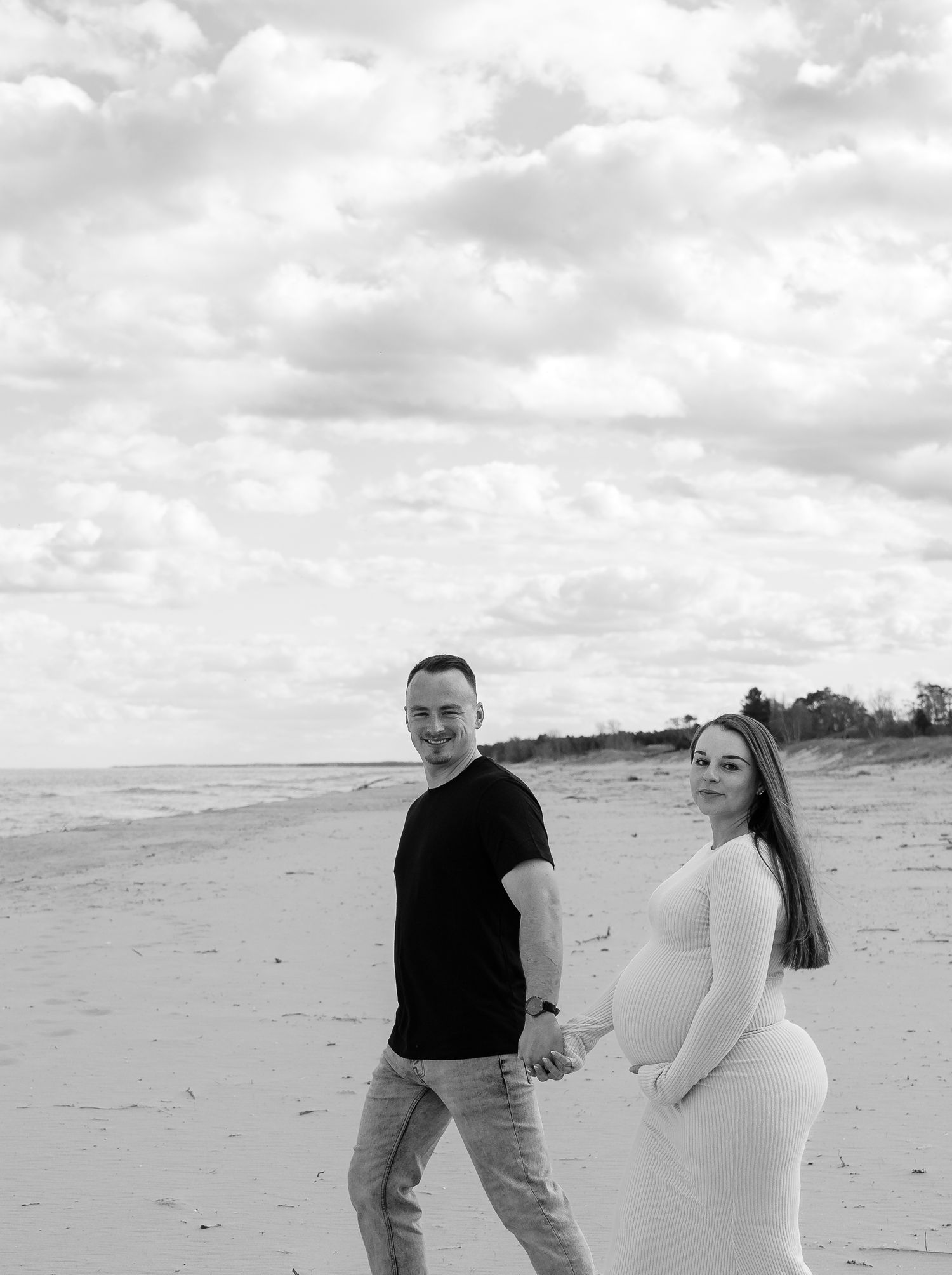 Black and white photo of a pregnant couple holding hands on a beach, smiling, with cloudy skies behind them.