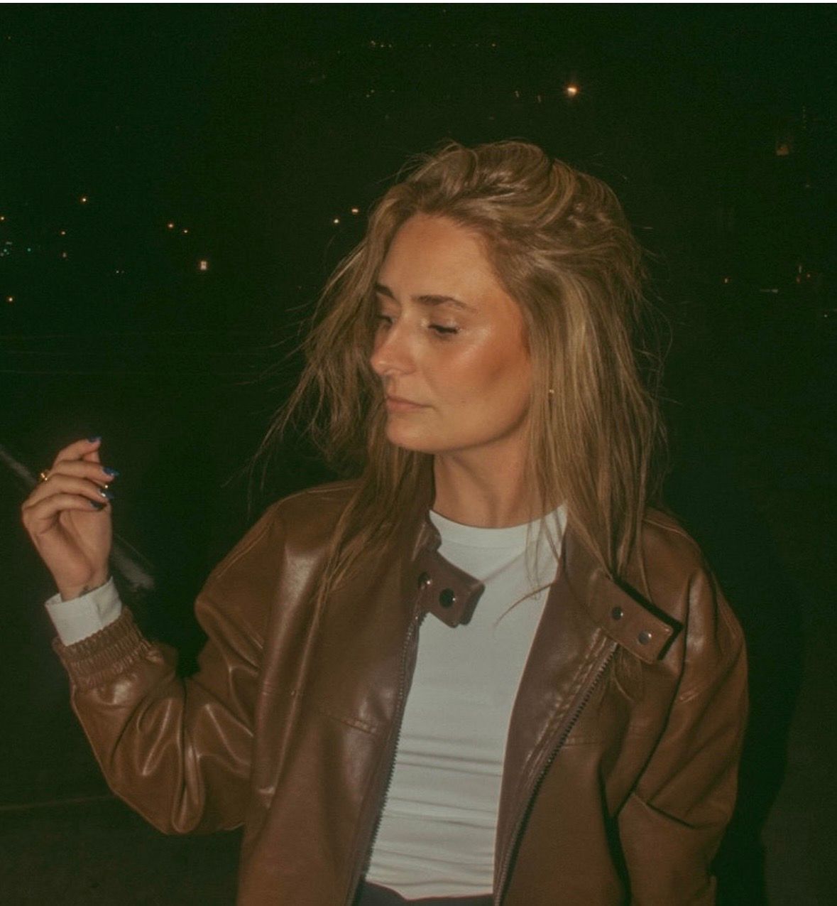 Woman in brown leather jacket and white top holding cigarette outdoors at night with tousled blonde hair.