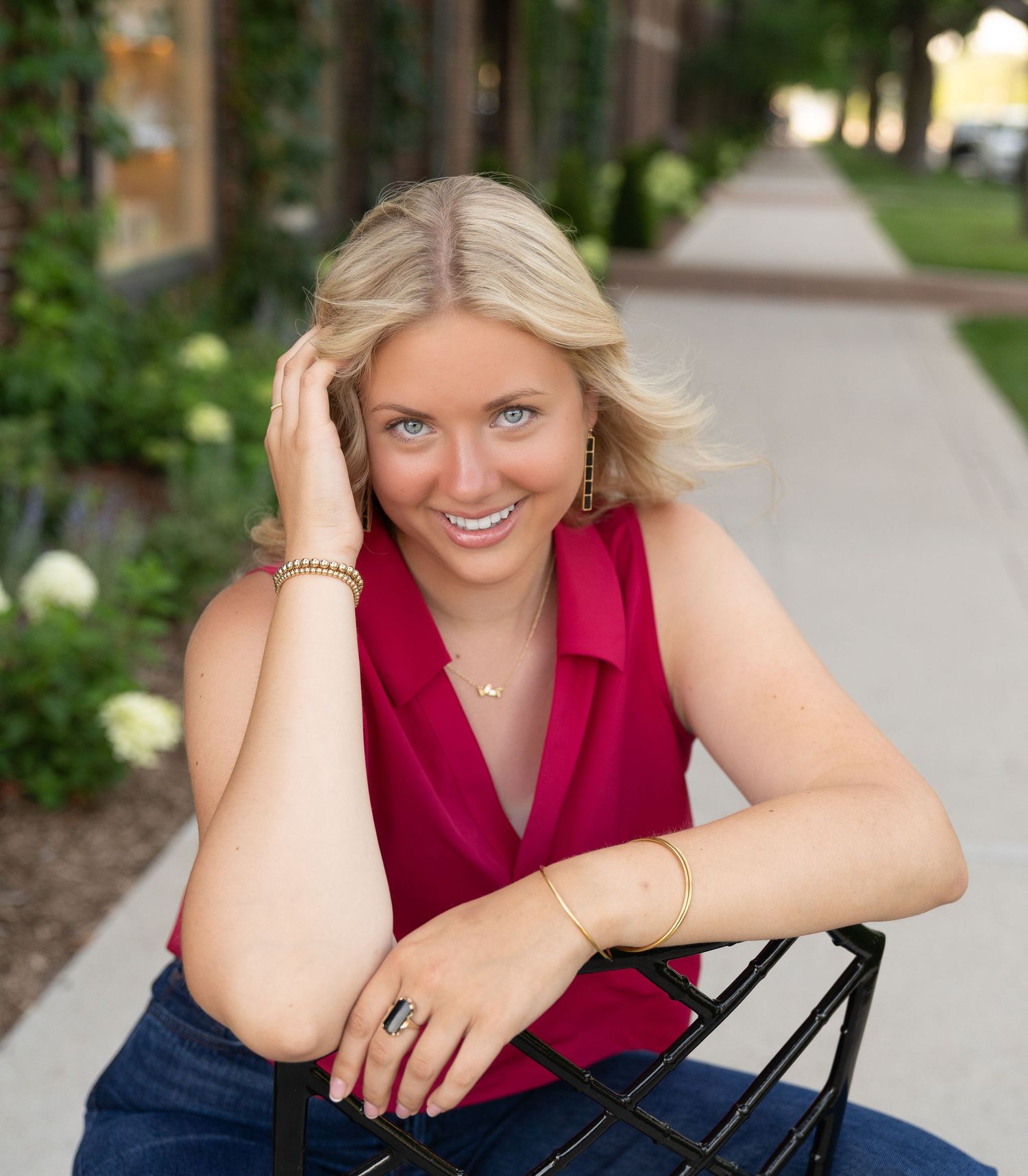 A smiling woman with blonde hair wearing a red top sits outdoors in a garden setting.