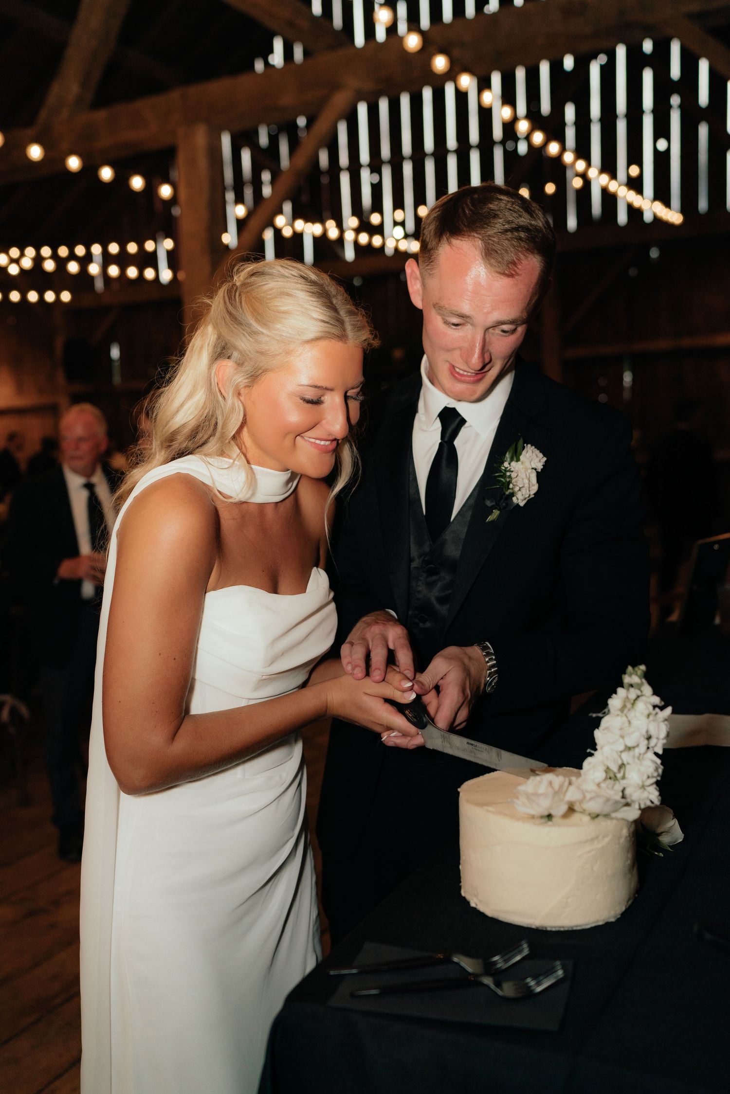 Bride and groom cutting their white wedding cake inside a barn reception venue with string lights.