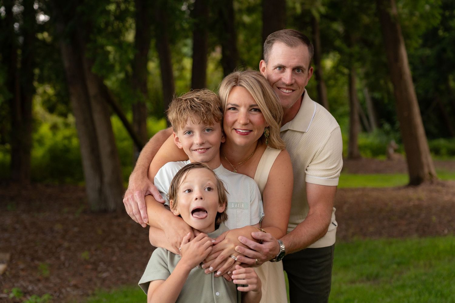 A happy family of four smiling and embracing in a wooded outdoor setting.