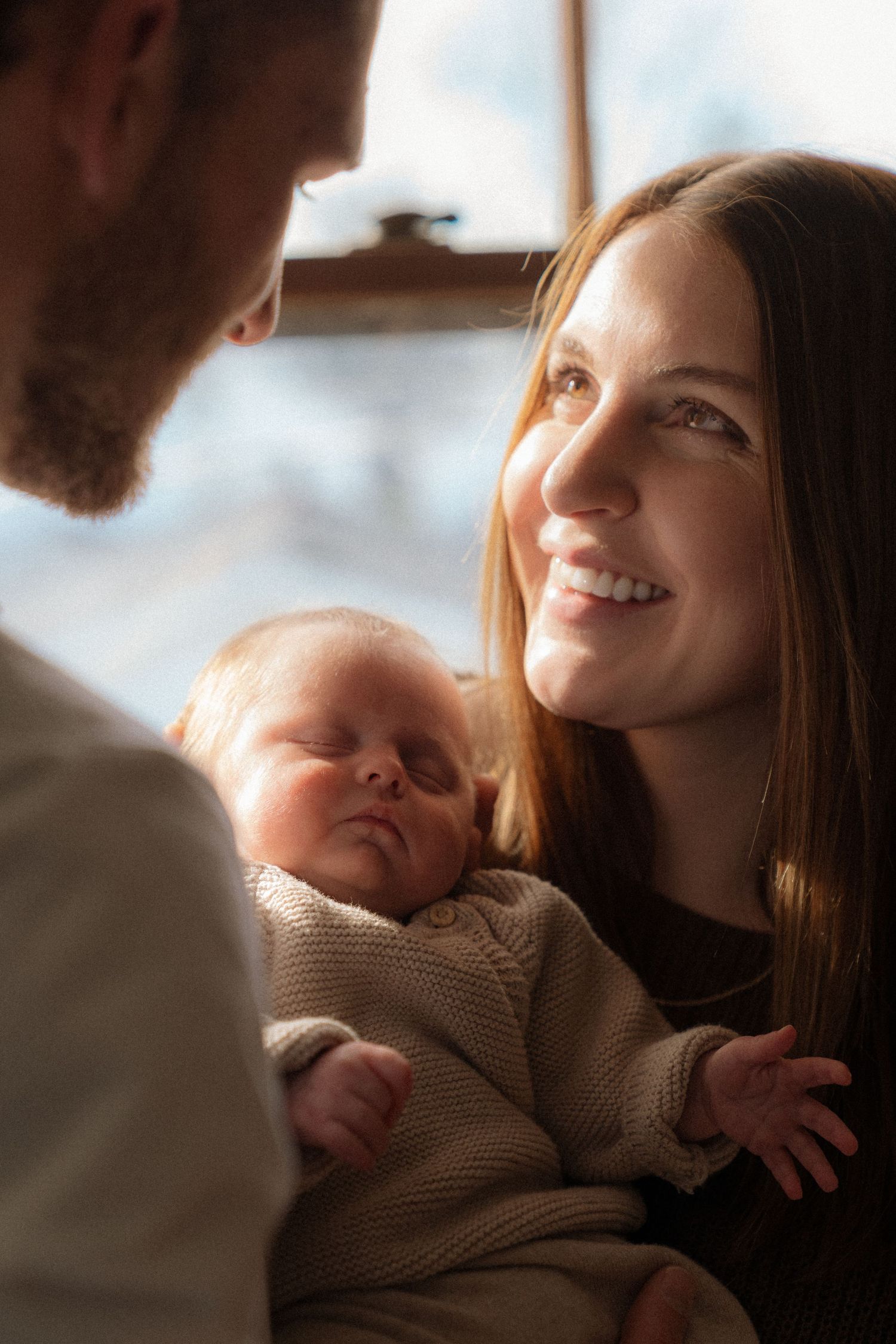 A smiling woman holds a sleeping newborn baby in her arms as she looks at the man in front of her.