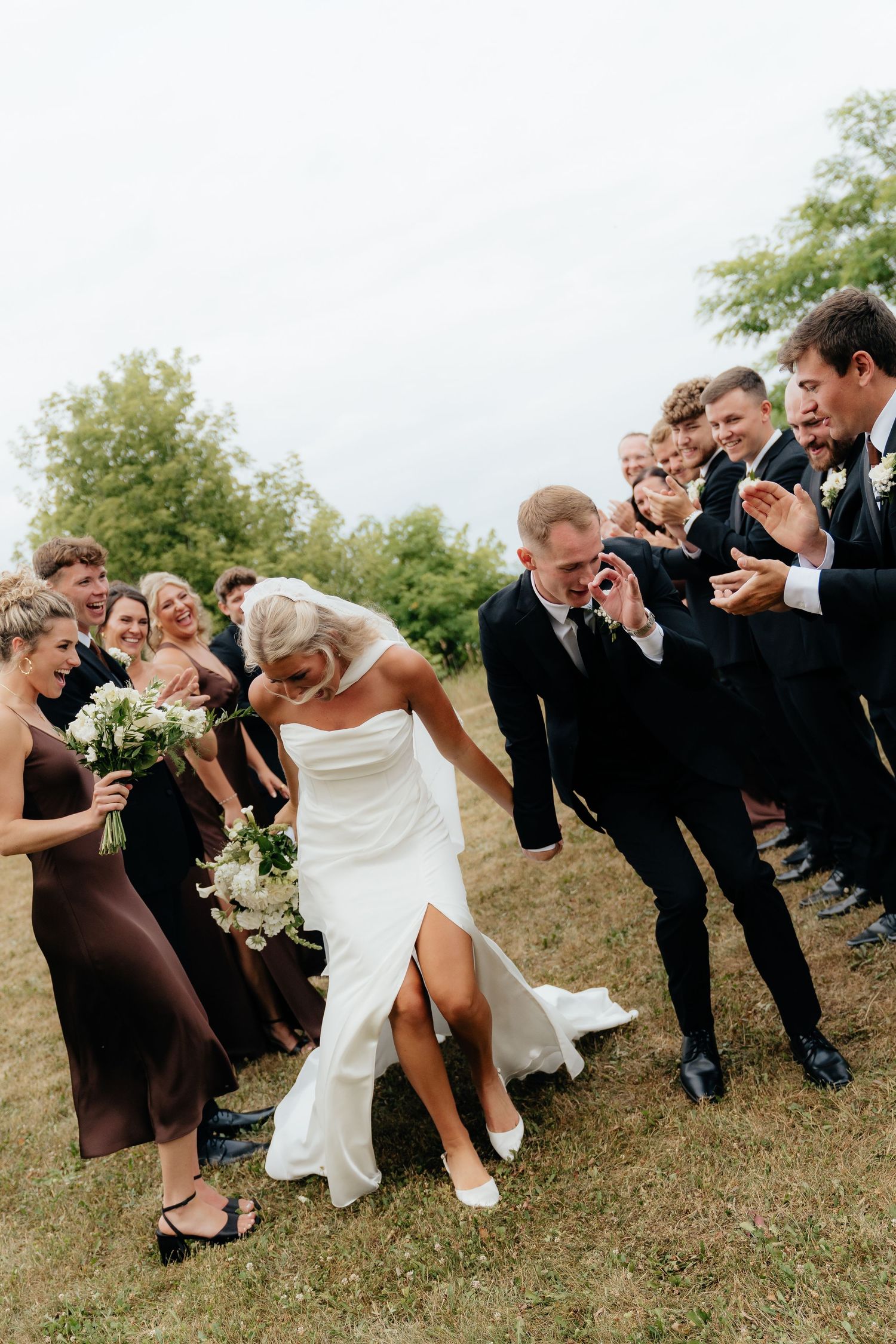 Bride and groom laugh joyfully as wedding party cheers them on outdoors on a green lawn.