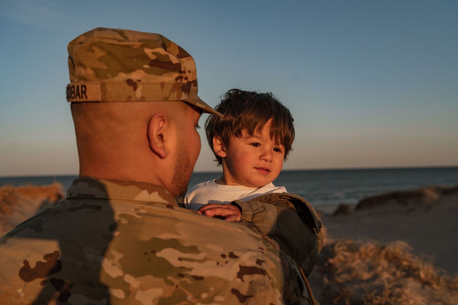 A soldier in camouflage uniform holds a young child on a beach at sunset.