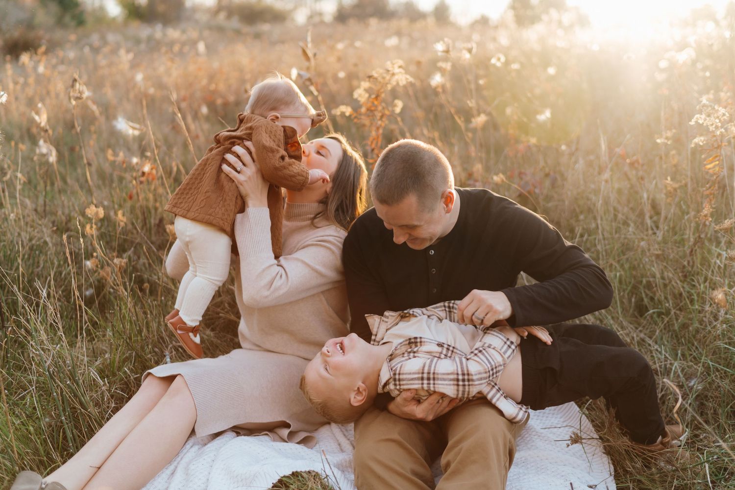 Family laughing together on a blanket in a sunlit golden field during a warm autumn outdoor photo session.