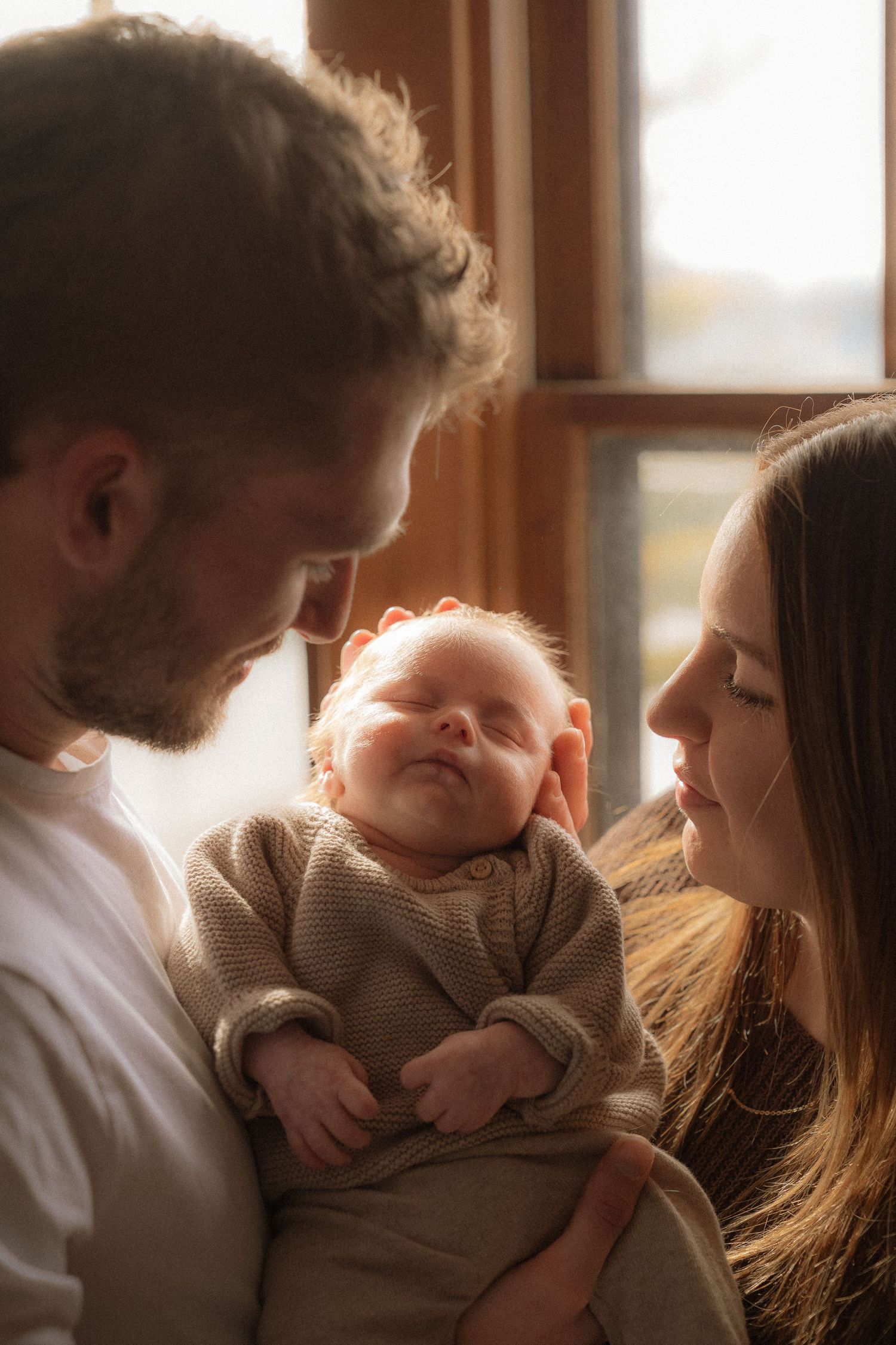 A father and mother are holding their newborn baby in a cozy, warm-lit room.