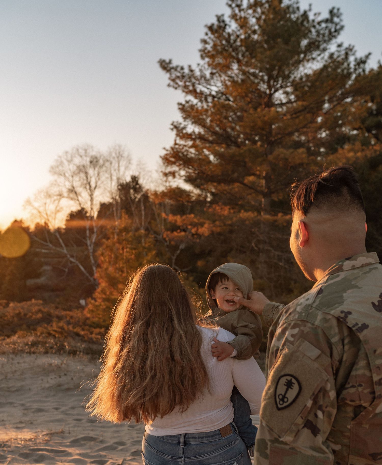 A soldier in camouflage uniform stands with a woman and baby at golden hour near pine trees.