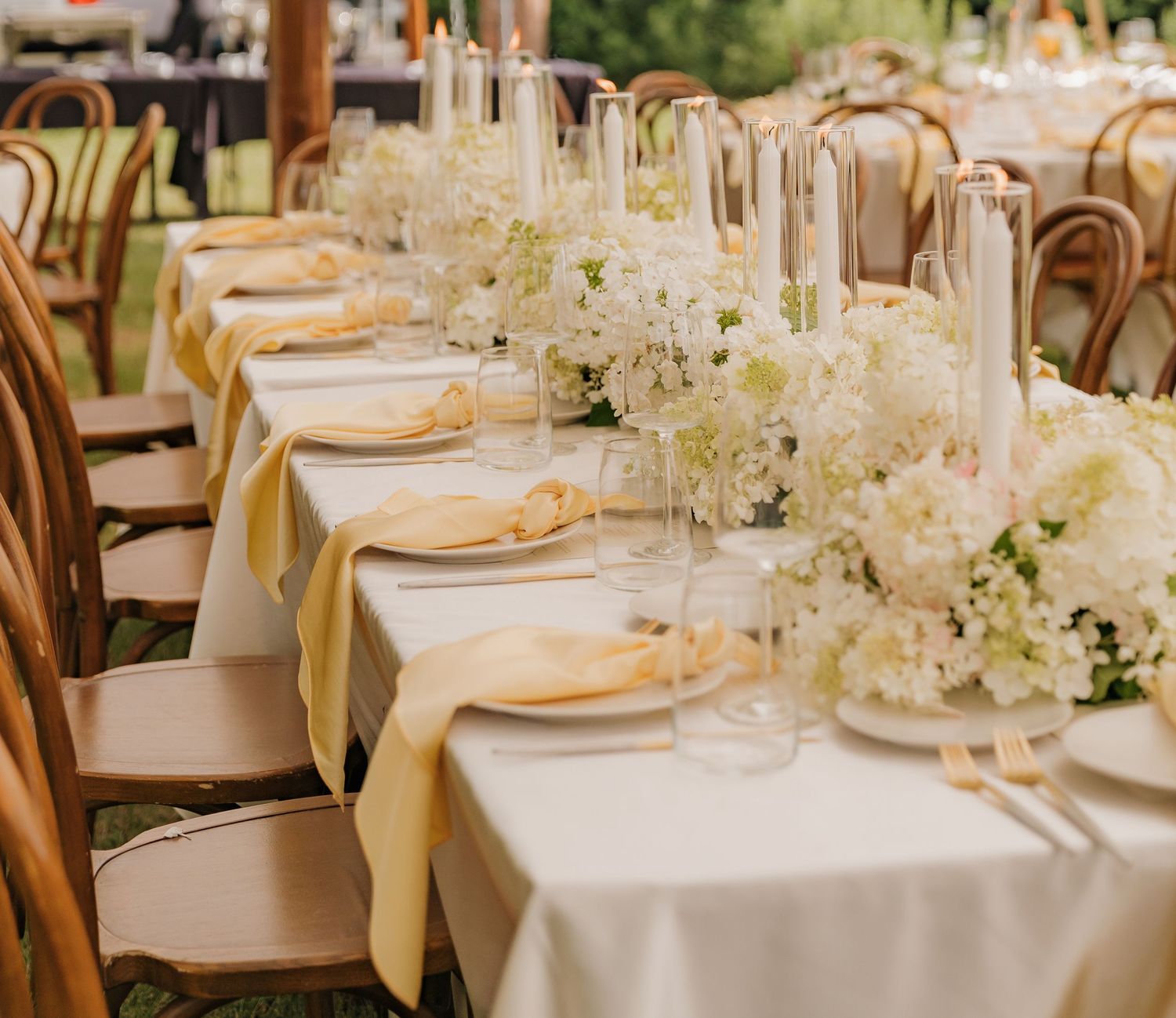 A formal wedding reception table with white floral centerpieces and golden linens set up in an outdoor setting.