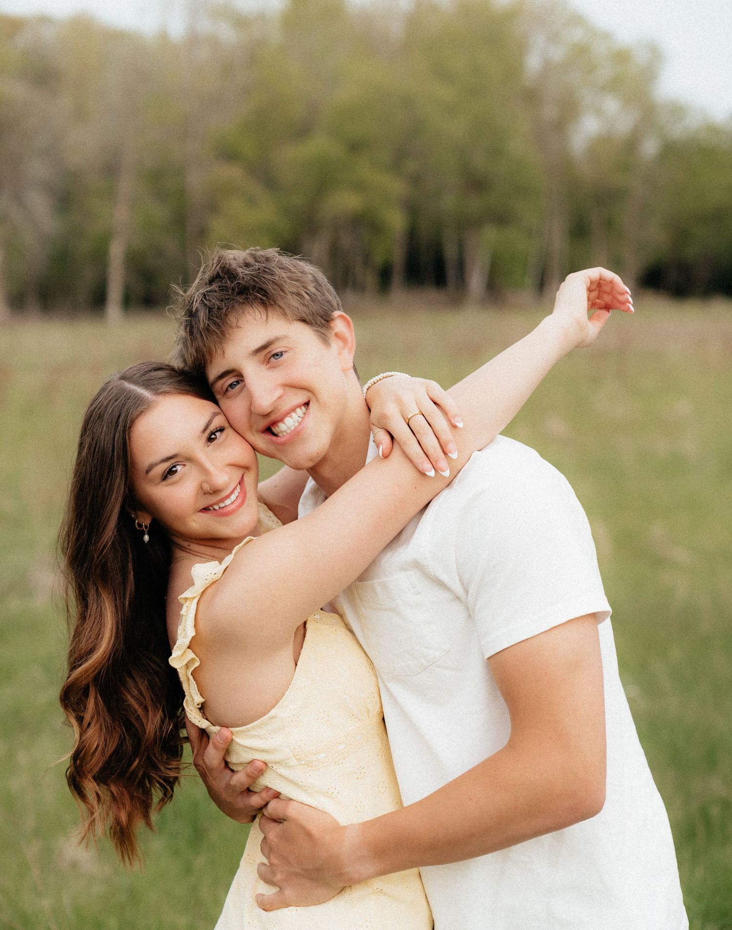 A young couple embracing and smiling outdoors in a green field, wearing casual summer clothing in a romantic portrait.